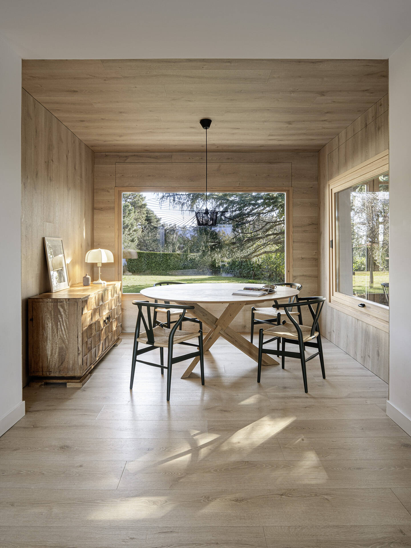 Warm timber wraps the walls and ceiling of this Costa Brava dining room, creating a cosy contrast to the home's white interiors. A rounded timber table, Carl Hansen chairs and large garden-facing windows complete the space.