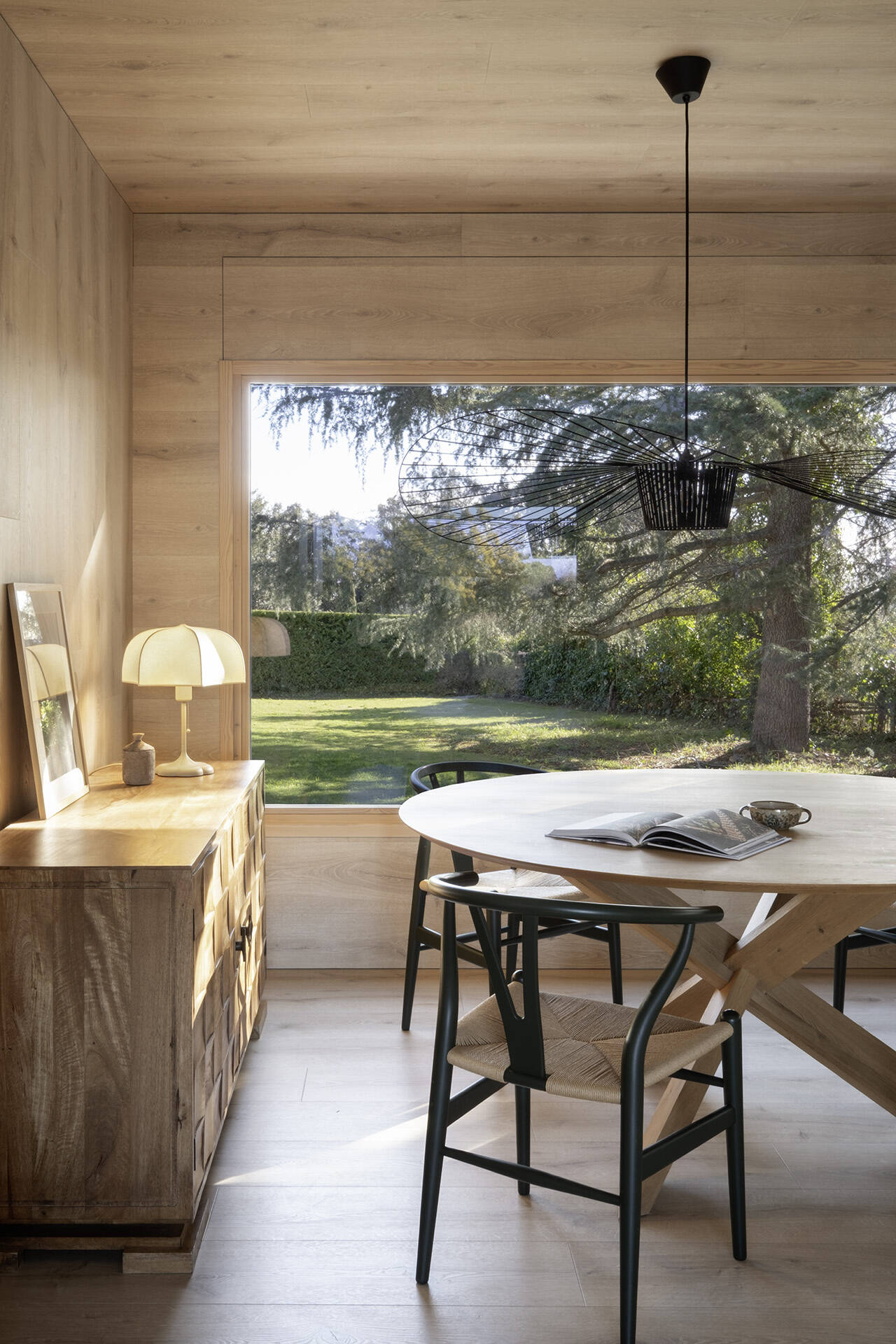 Warm timber wraps the walls and ceiling of this Costa Brava dining room, creating a cosy contrast to the home's white interiors. A rounded timber table, Carl Hansen chairs and large garden-facing windows complete the space.
