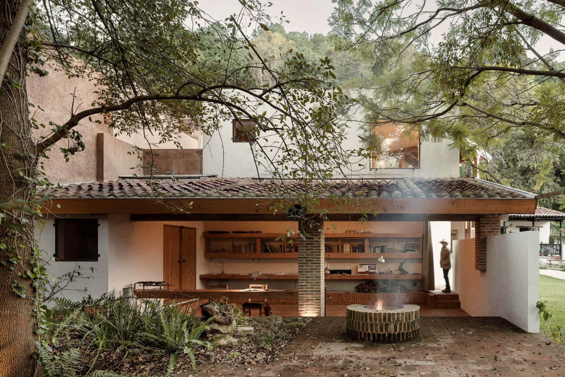 Step into Casa Amona’s courtyard in Chiapas, México, where an existing ash tree shades a brick fire pit and a covered workspace opens onto the planting.