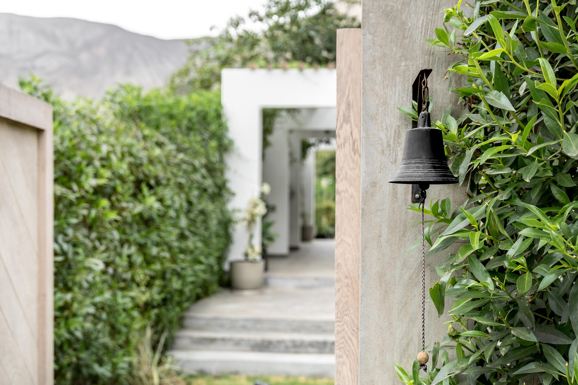 Step inside Casa Berna through a shaded walkway of white frames and timber poles, with lime-washed brick, lush planting and a garden view beyond.