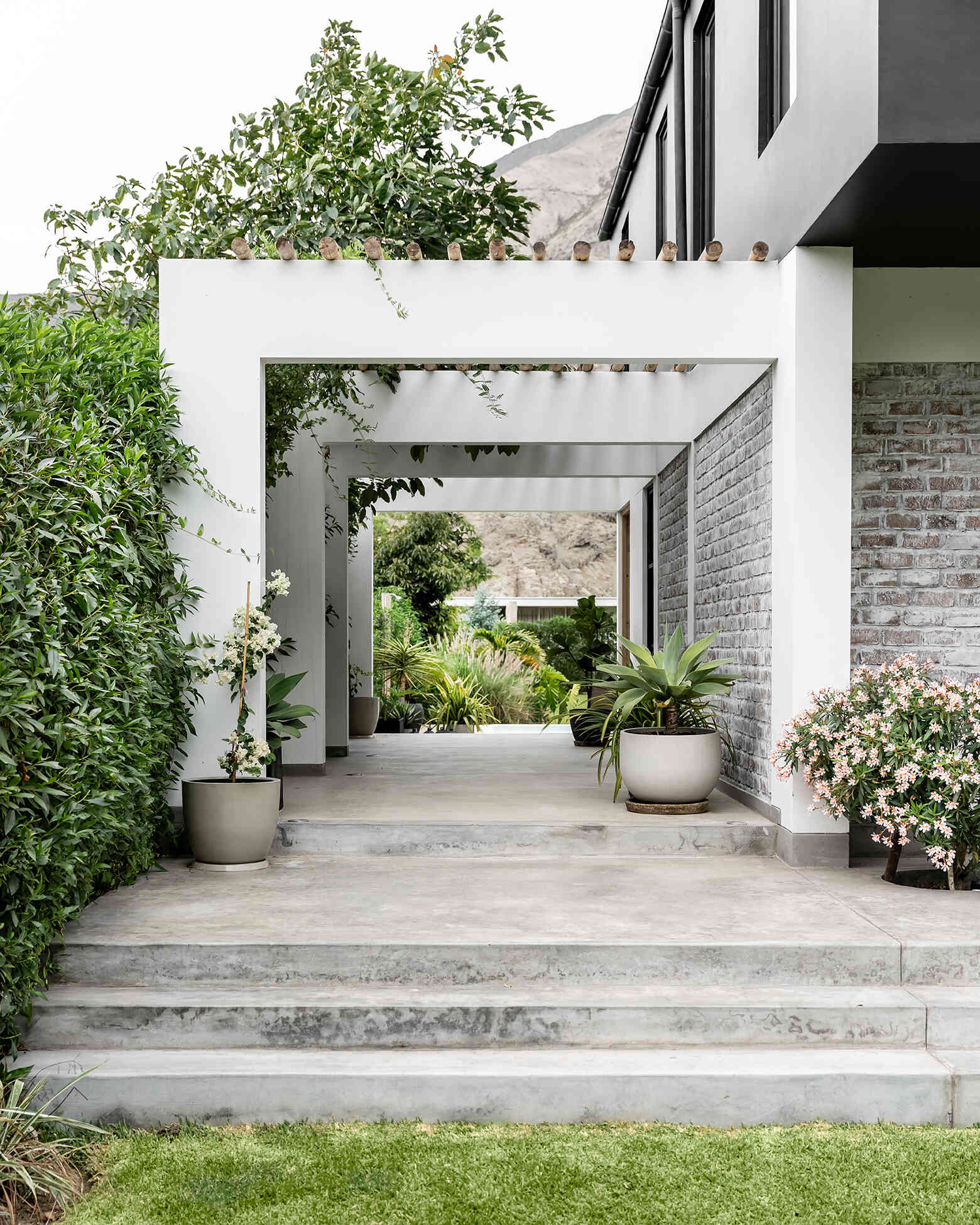 Step inside Casa Berna through a shaded walkway of white frames and timber poles, with lime-washed brick, lush planting and a garden view beyond.
