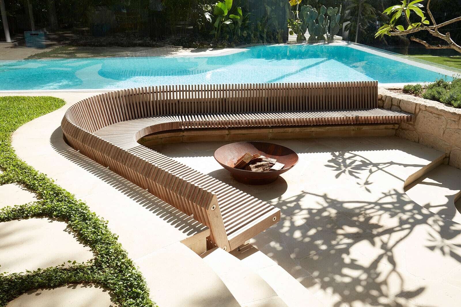 A large curved timber bench curves around a fire bowl in the backyard, creating a distinctive sunken seating area surrounded by lush planting.