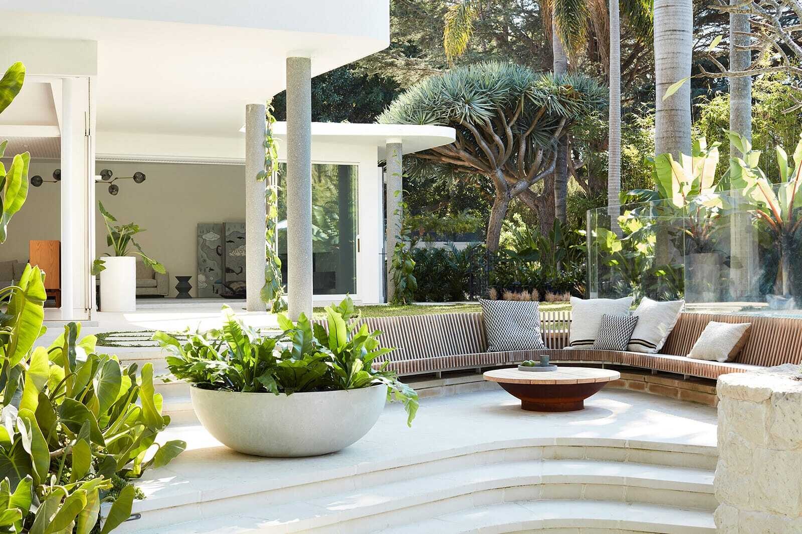 A large curved timber bench curves around a fire bowl in the backyard, creating a distinctive sunken seating area surrounded by lush planting.