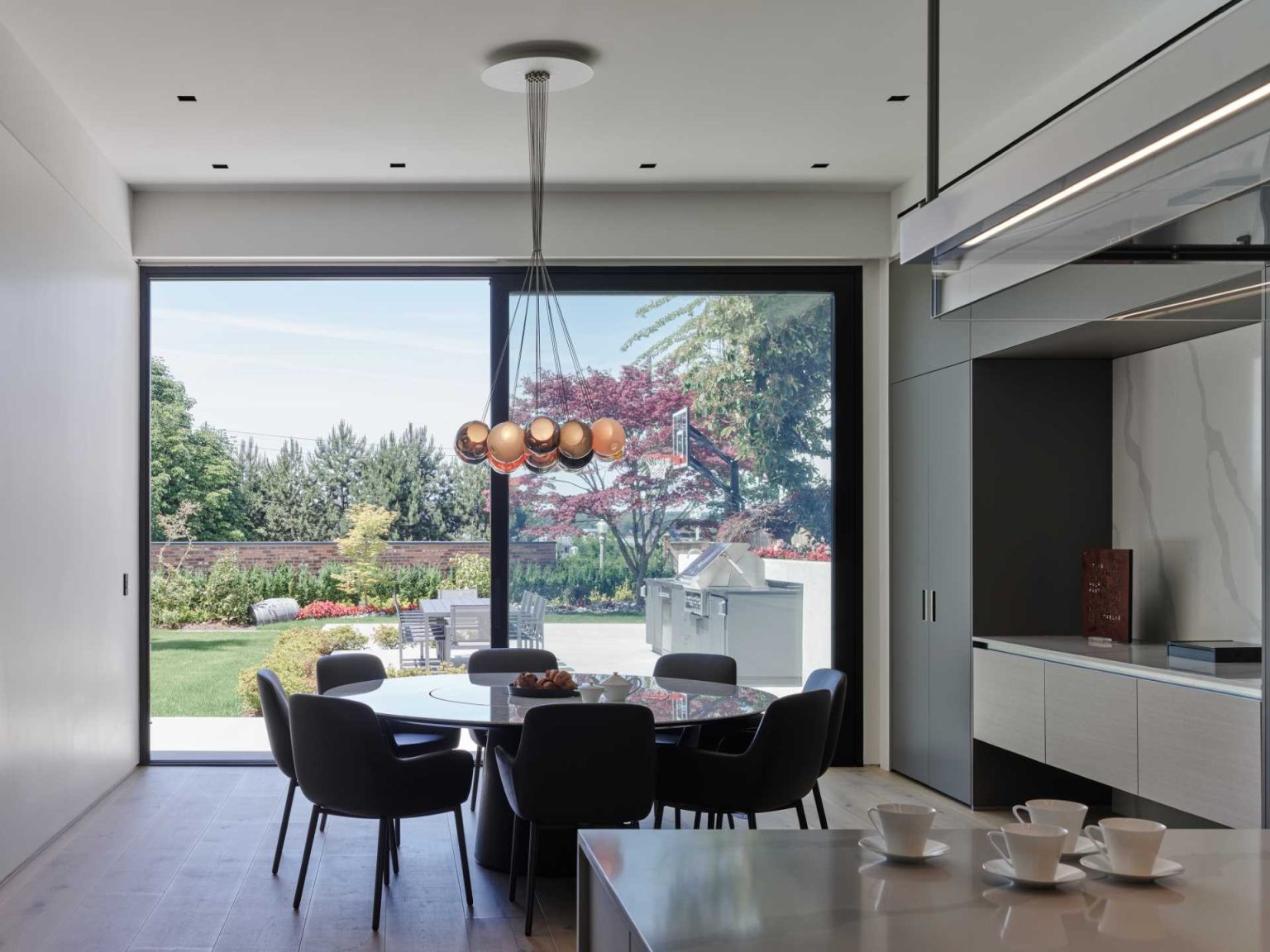 Grey cabinetry stretches along the kitchen and dining wall of Nanton House, contrasting with warm wood floors and millwork, while an island provides bar seating and garden views.