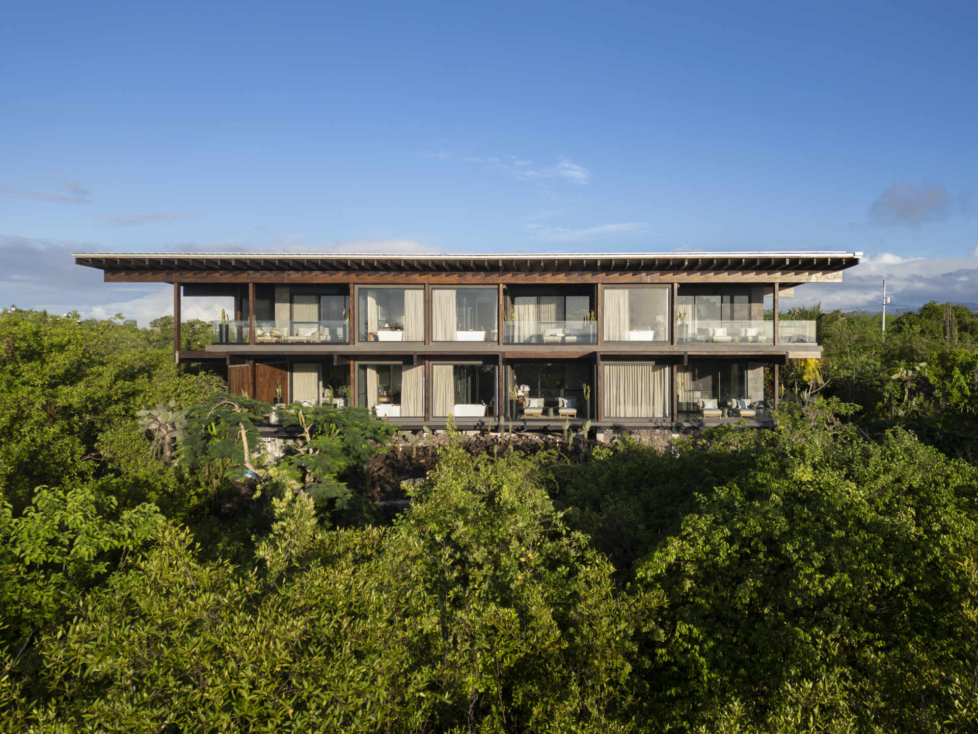 Explore the Finch Bay Galápagos Hotel expansion near Puerto Ayora, Ecuador, by Alfredo Ribadeneira Arquitectos. Prefabricated teak construction, deep roof overhangs, glass-fronted terraces, and neutral guest spaces frame views of trees and volcanic rock.