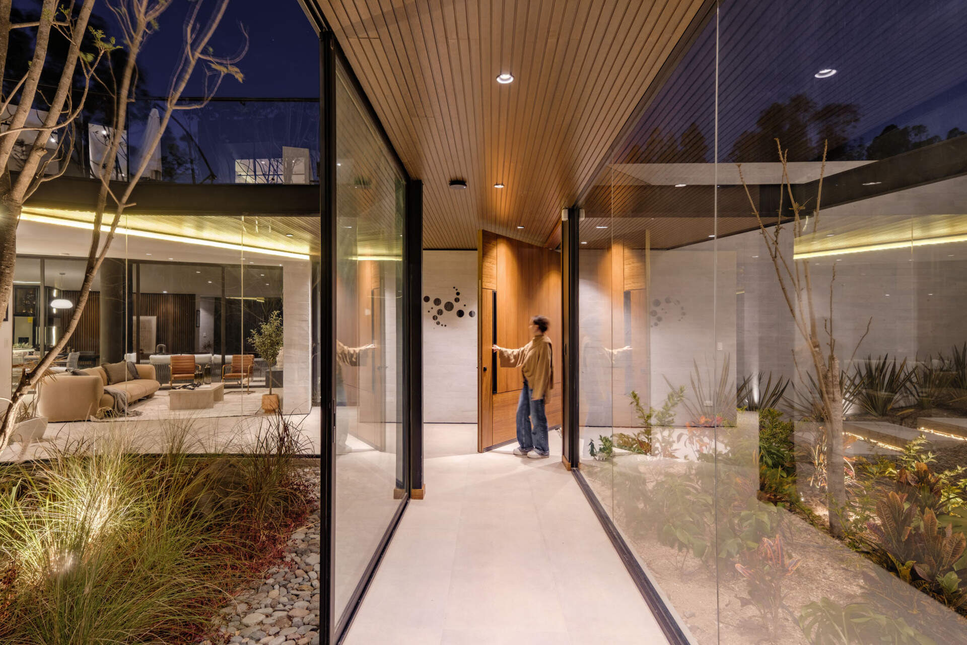 A glass hallway wraps around an open-air courtyard planted with a tall tree, ornamental grasses, and low red foliage. Timber ceilings, pale tiled floors, and upper walkways with glass balustrades bring garden views into the circulation spaces of this contemporary Ecuadorian home.
