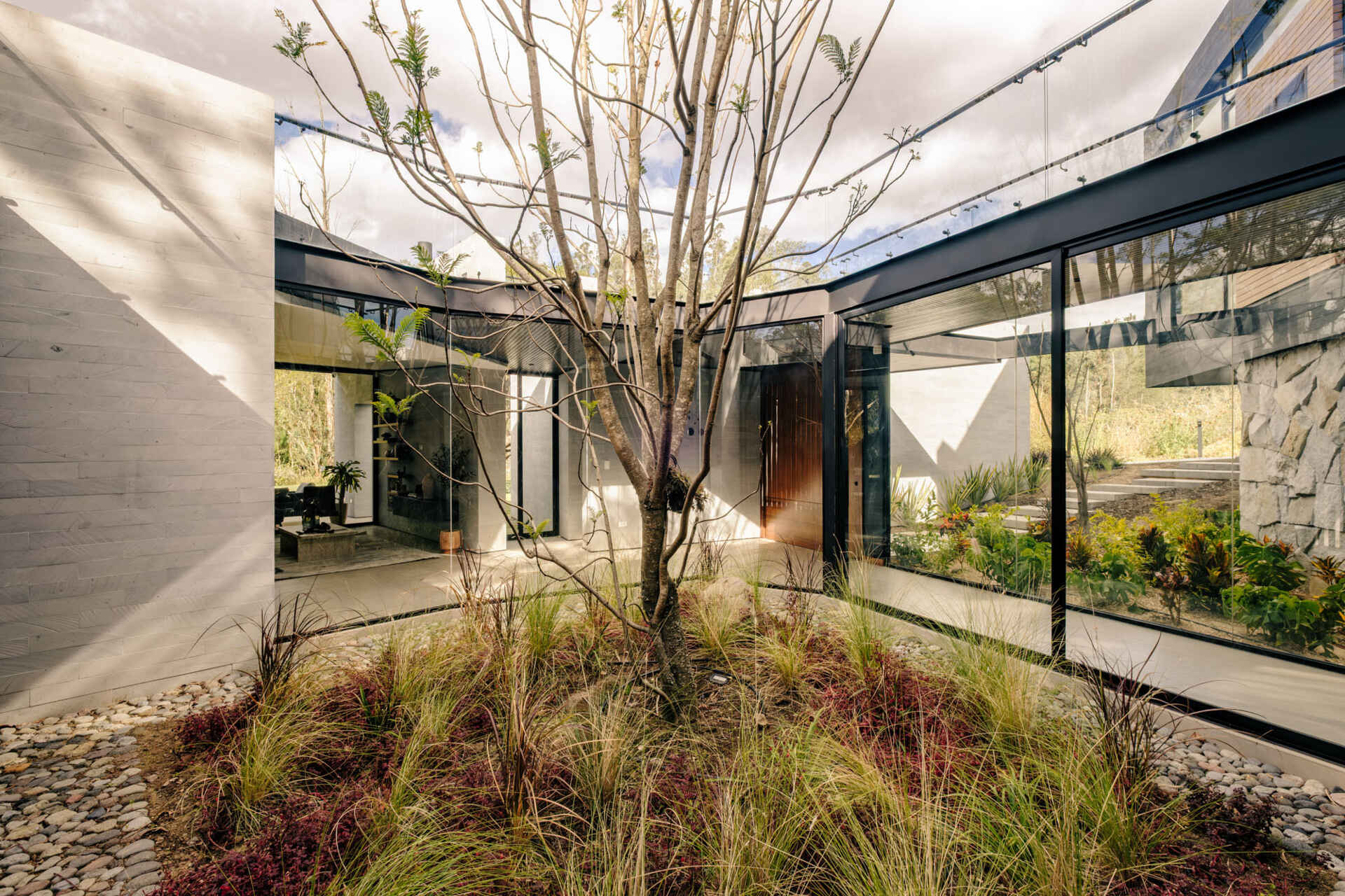 A glass hallway wraps around an open-air courtyard planted with a tall tree, ornamental grasses, and low red foliage. Timber ceilings, pale tiled floors, and upper walkways with glass balustrades bring garden views into the circulation spaces of this contemporary Ecuadorian home.