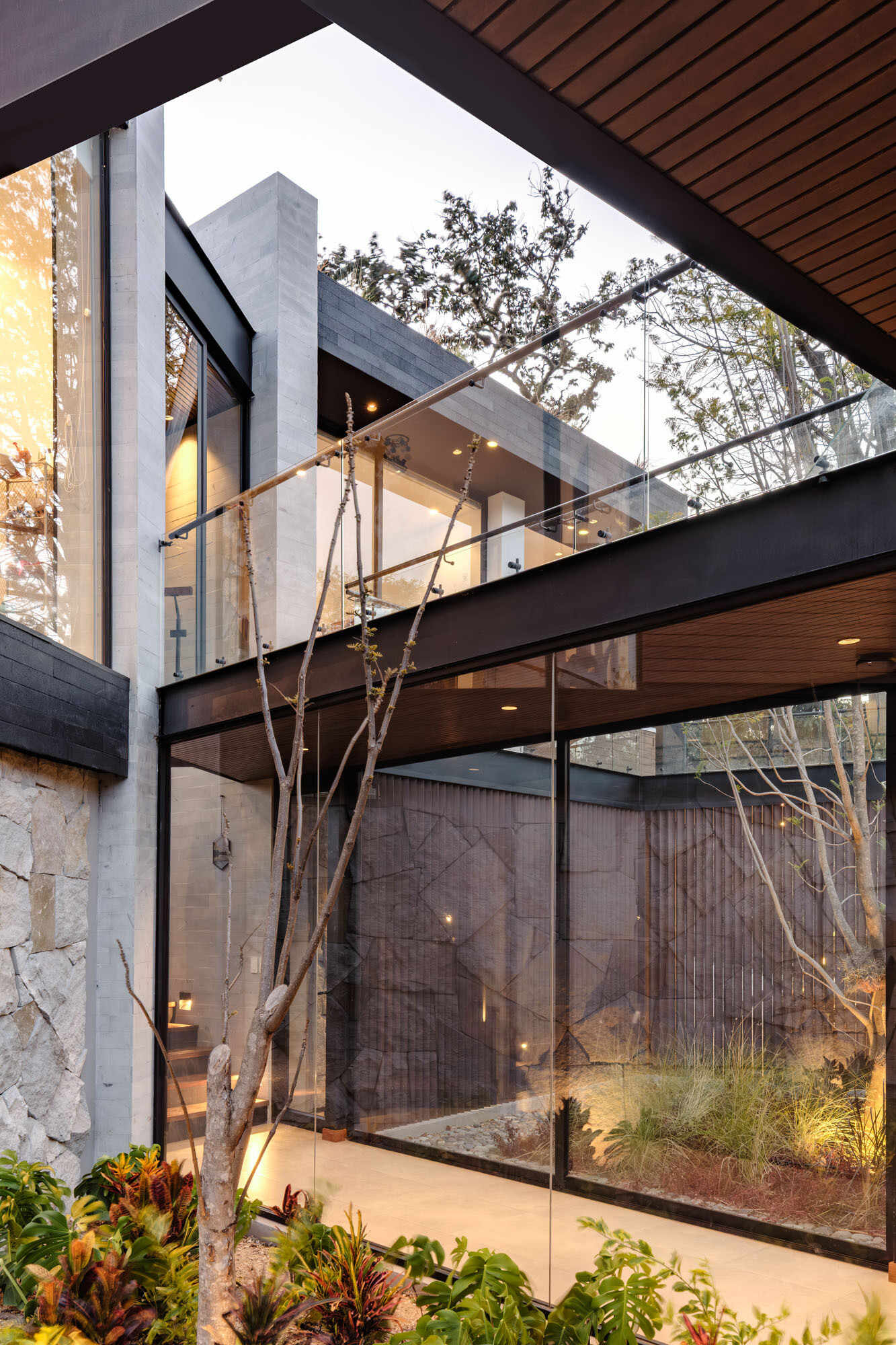 A glass hallway wraps around an open-air courtyard planted with a tall tree, ornamental grasses, and low red foliage. Timber ceilings, pale tiled floors, and upper walkways with glass balustrades bring garden views into the circulation spaces of this contemporary Ecuadorian home.