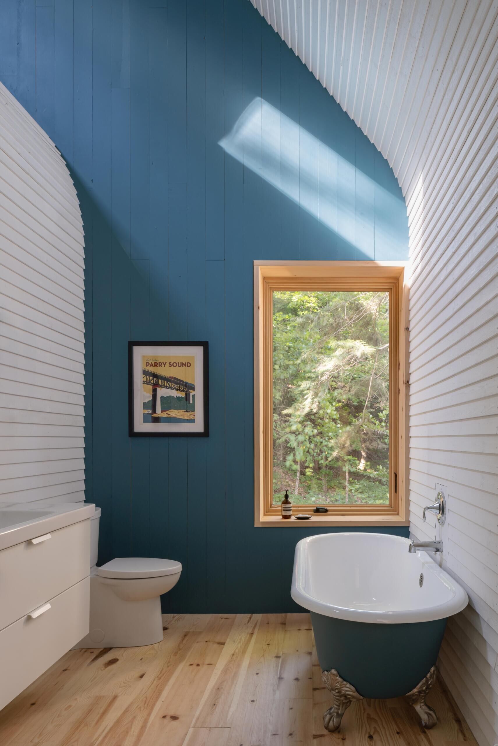 A modern cottage bathroom featuring a blue painted gable wall, reclaimed bathtub, curved timber ceiling, and a warm minimalist design overlooking the forest.