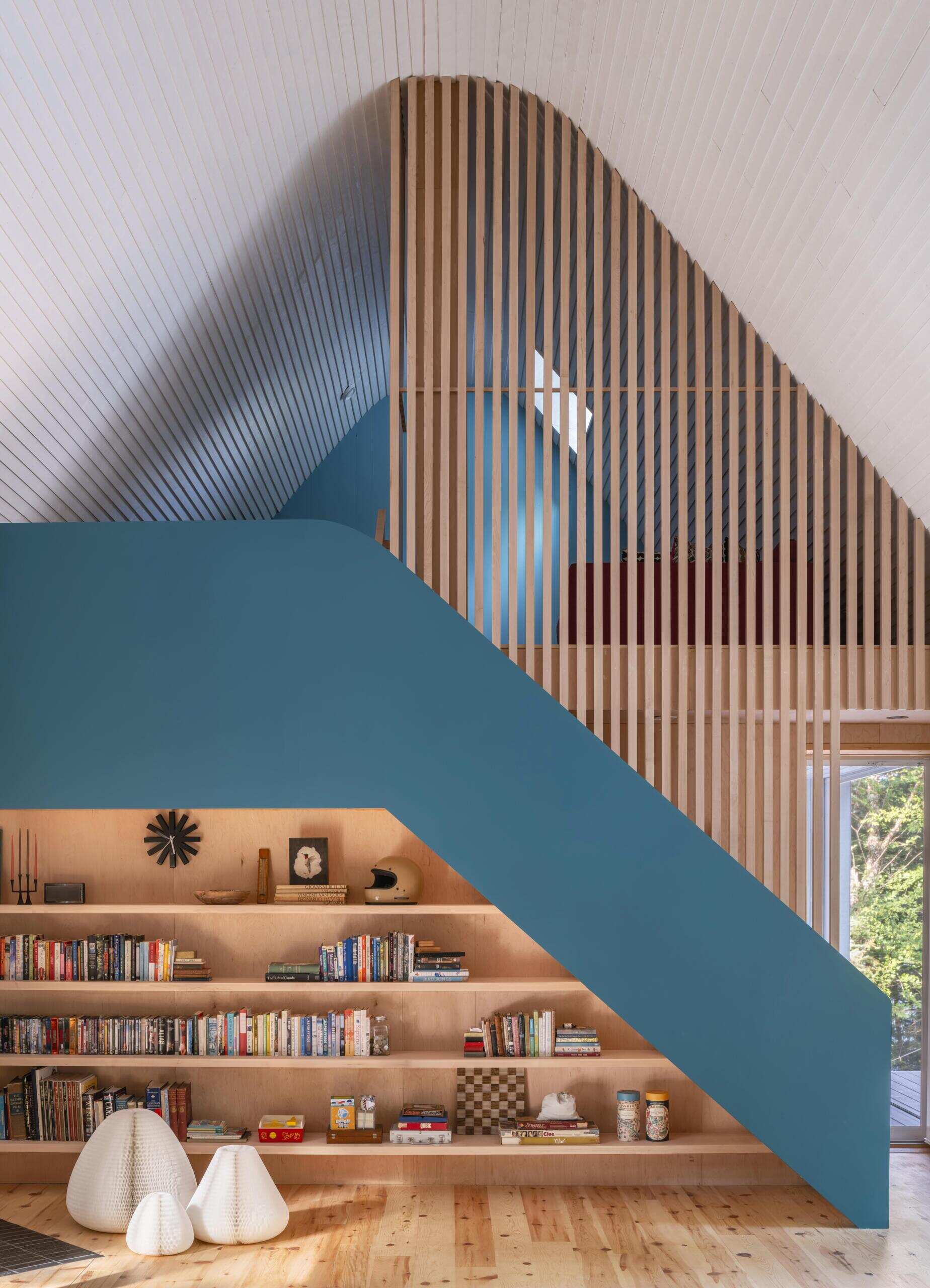 A modern cottage living room with built-in timber shelving, a sculptural blue staircase, maple slat loft screen, and warm minimalist interior details.