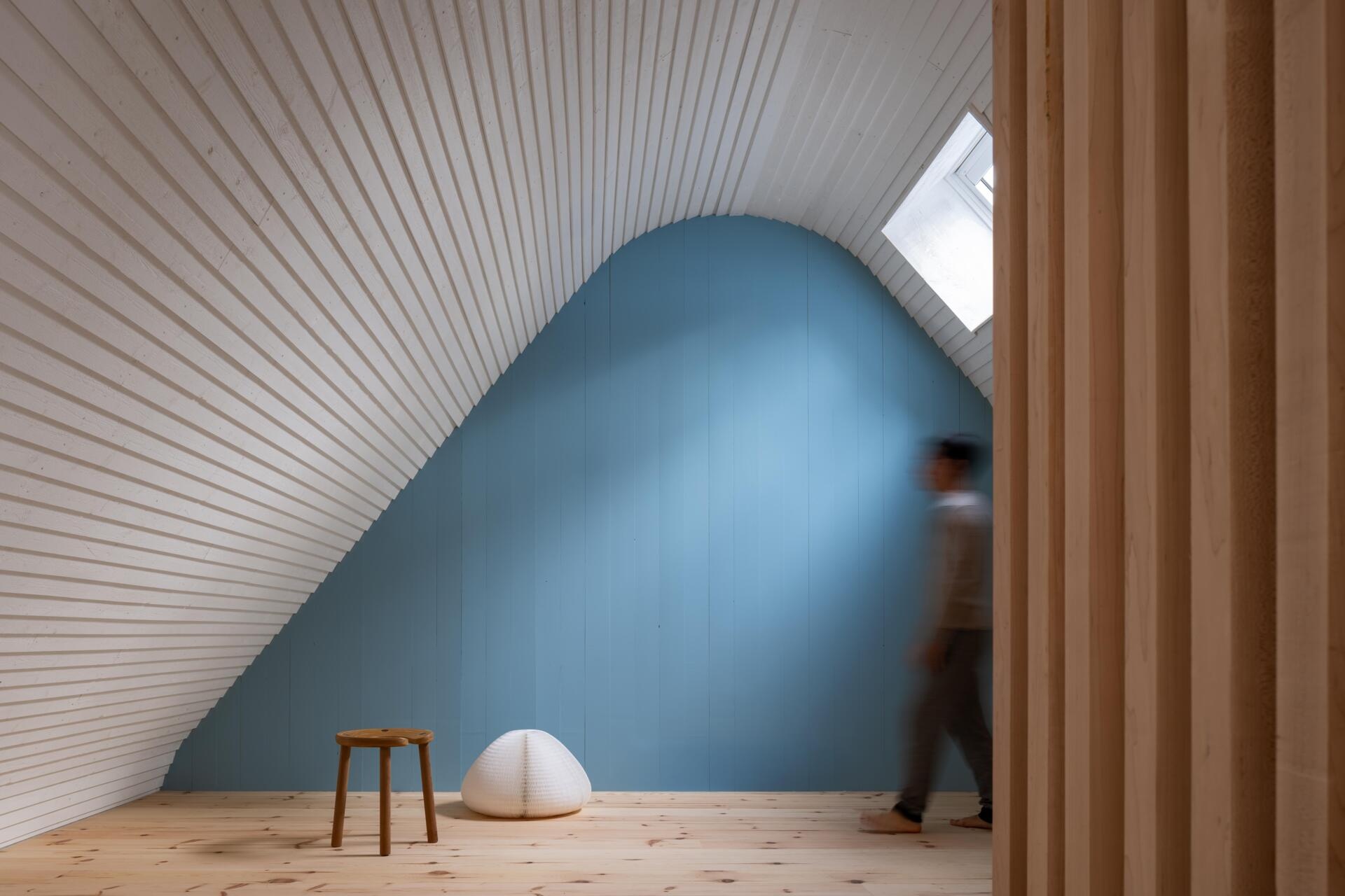 A modern cottage loft featuring a curved white timber ceiling, blue painted gable wall, maple slat screen, and sculptural staircase within a warm minimalist interior.