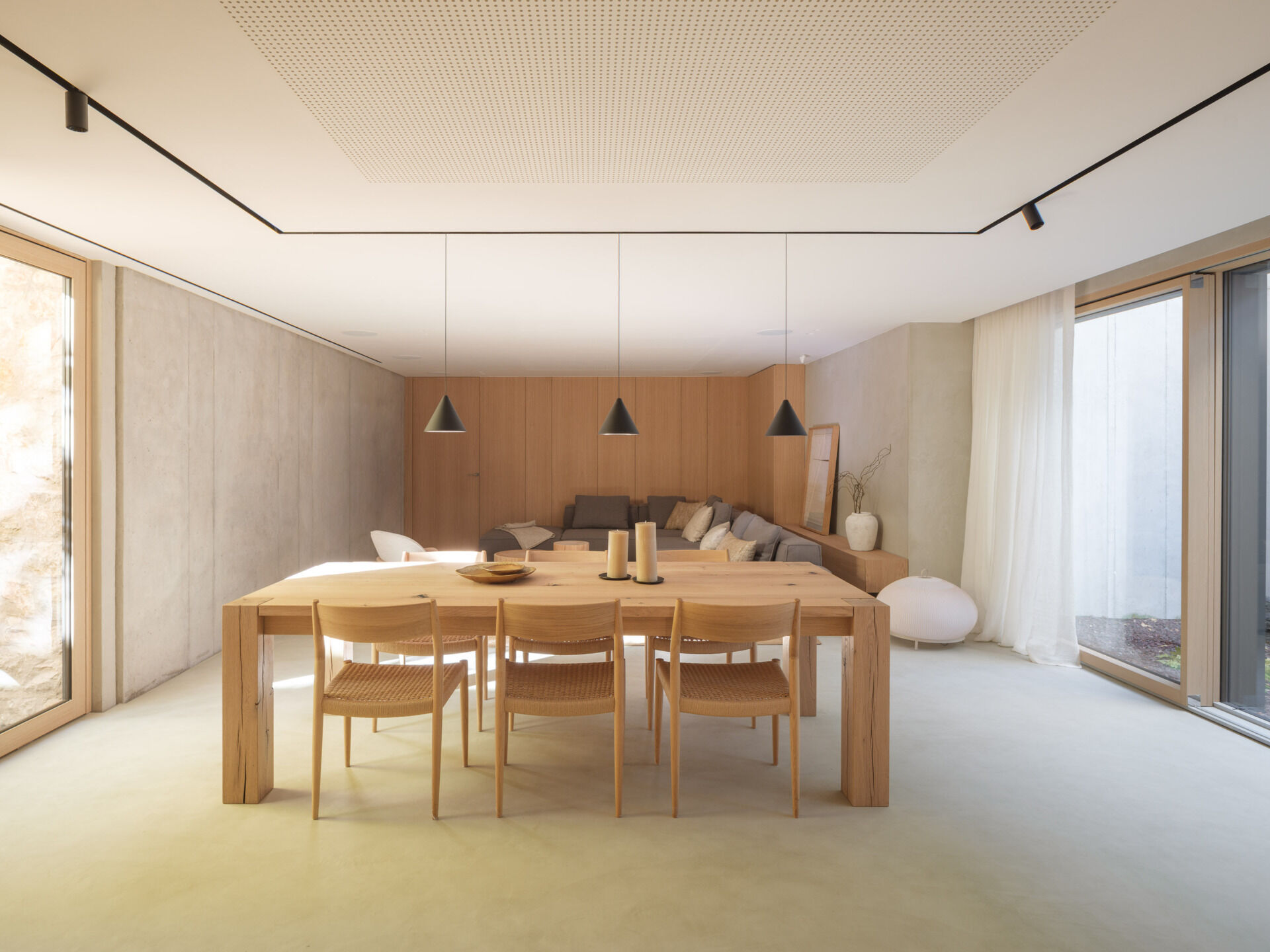 Sunken courtyards bring natural light into this basement living and dining area at House 2194 in Olot, Spain. Exposed concrete, timber joinery, woven dining chairs, and a grey sectional create a relaxed space for gathering.