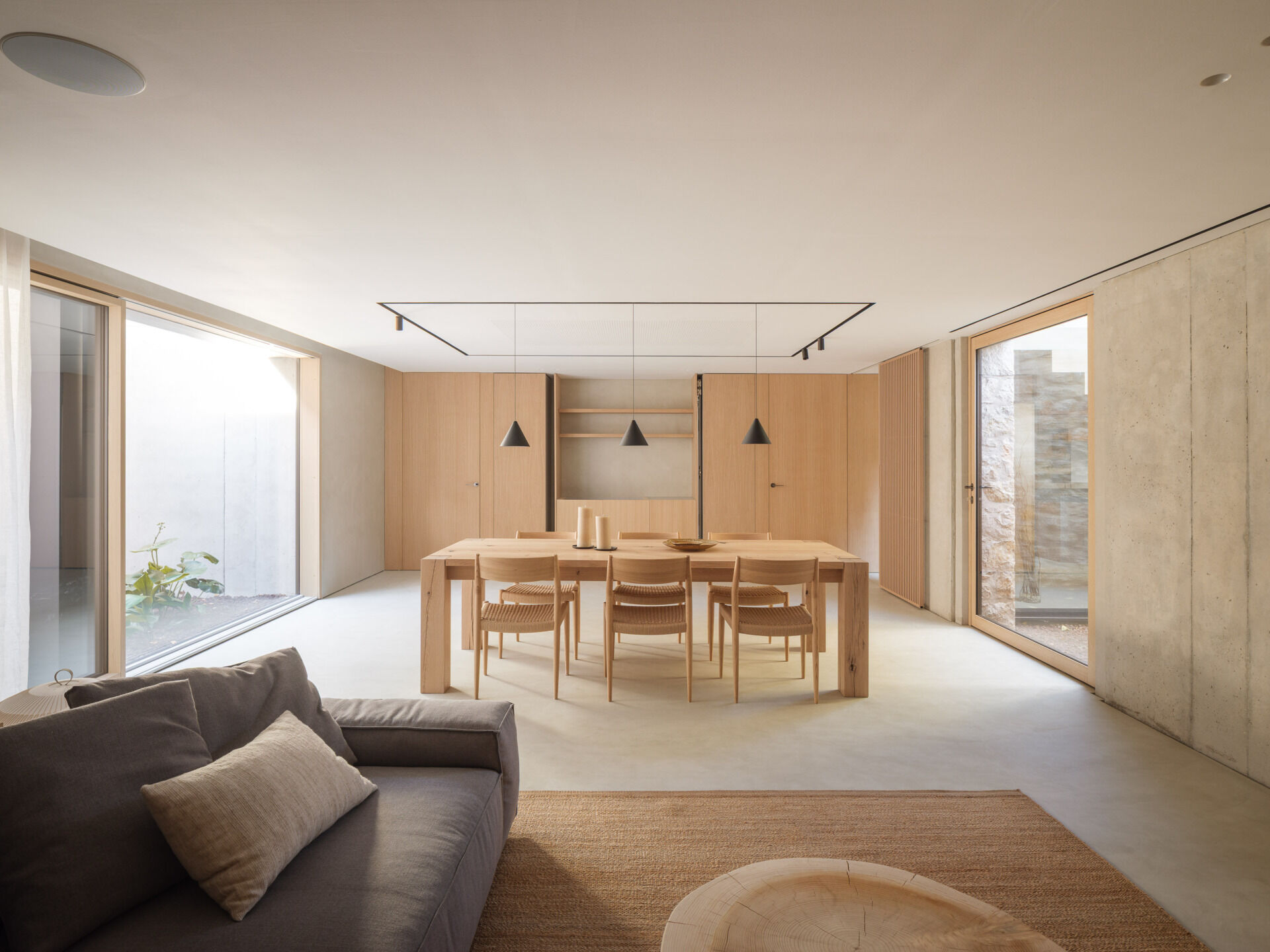 Sunken courtyards bring natural light into this basement living and dining area at House 2194 in Olot, Spain. Exposed concrete, timber joinery, woven dining chairs, and a grey sectional create a relaxed space for gathering.