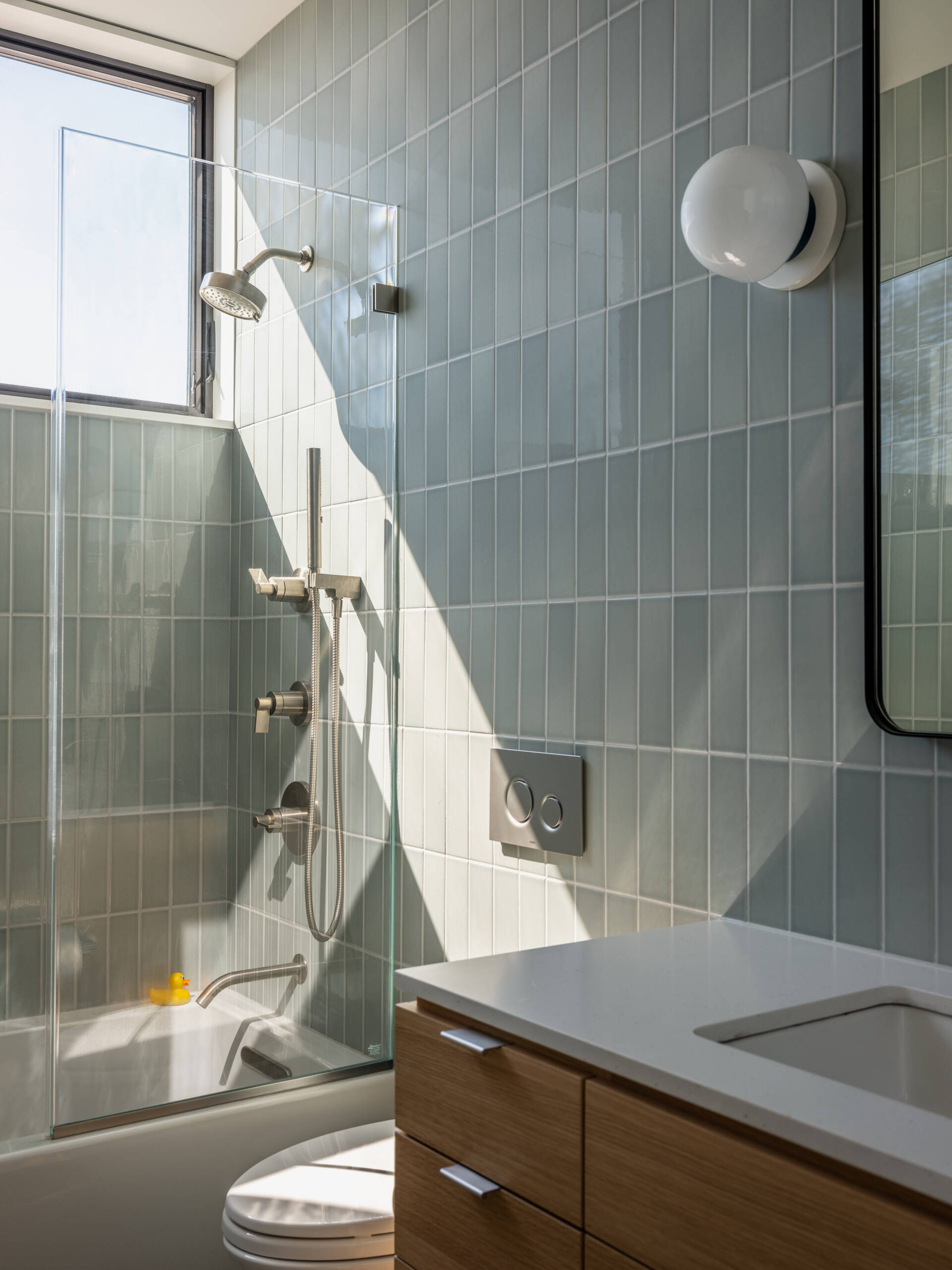 Pale green vertical tiles, a wood vanity, and a clear glass bath screen create a bright bathroom at Surf Road House. A high window brings in natural light, complemented by a white globe wall light and black-framed mirror.