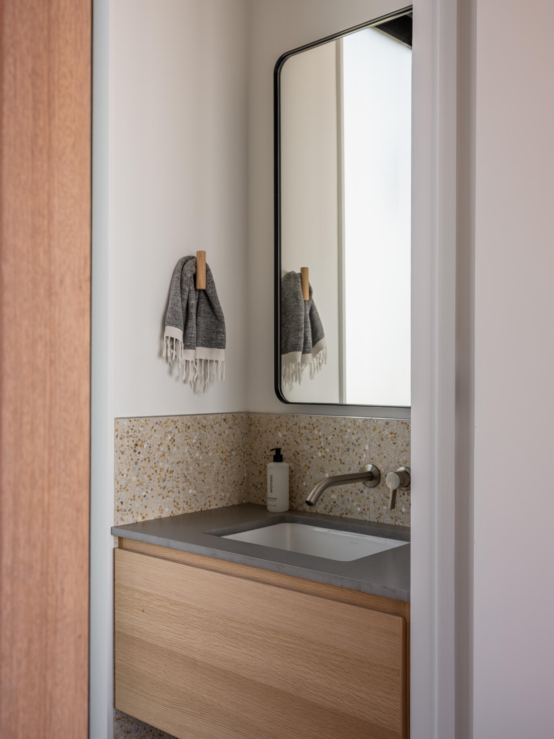 A compact wood vanity pairs a grey countertop with a speckled backsplash in Surf Road House. Wall-mounted tapware, a rounded black-framed mirror, and a wooden towel hook offer inspiration for a small bathroom or powder room.
