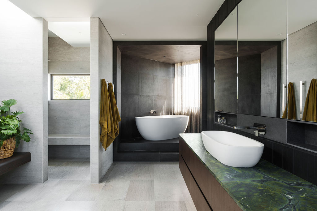 Inside the Mezzanine House bathroom, soft gray stone, dark surfaces, timber cabinetry and a green stone countertop create a calm contemporary space with a freestanding bathtub.