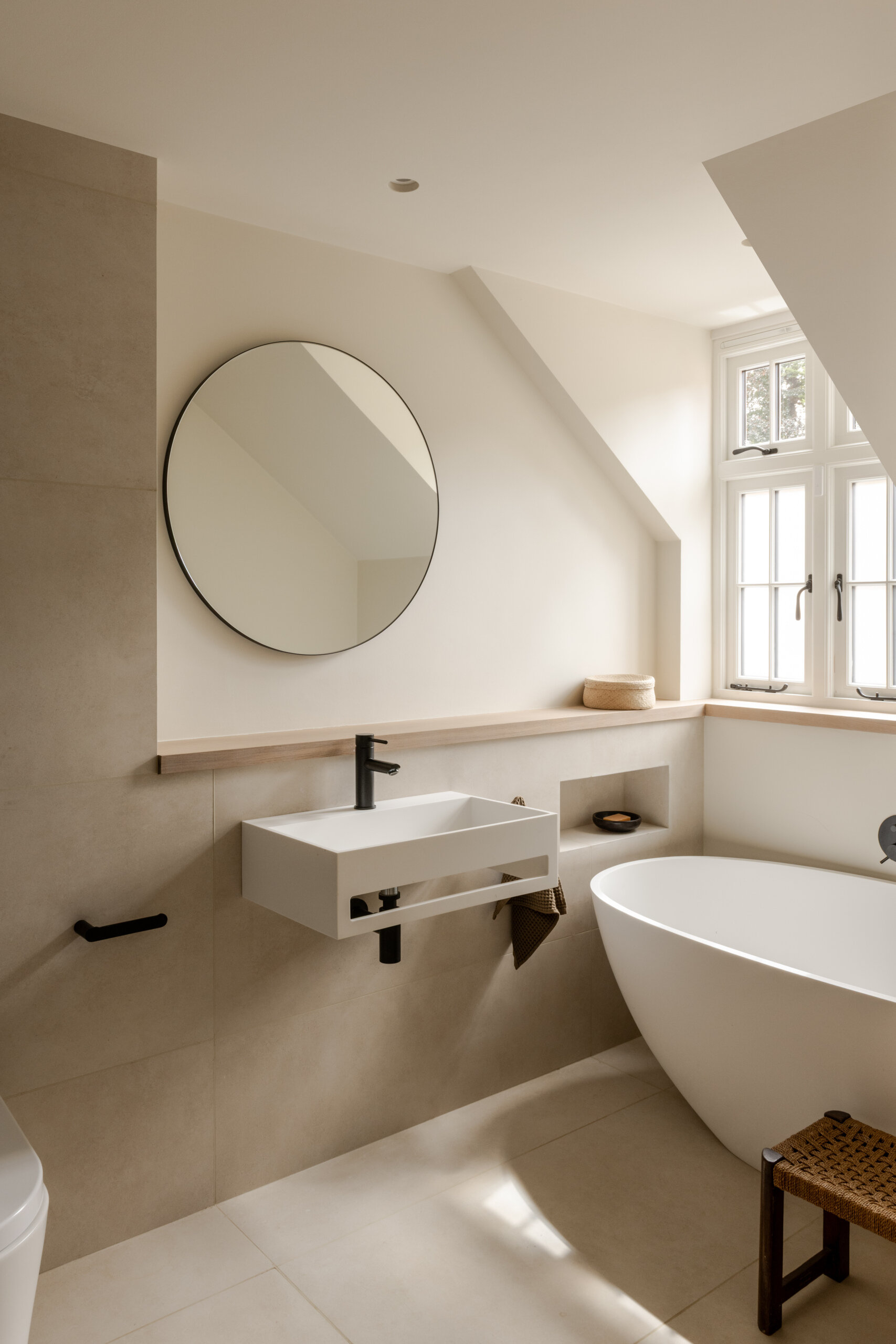 In this bathroom, a freestanding tub sits beneath a sloped ceiling, paired with warm white walls, natural wood details and slim black fixtures. A wall-mounted sink, round mirror and woven stool keep the compact room feeling open, while muted textiles add softness.