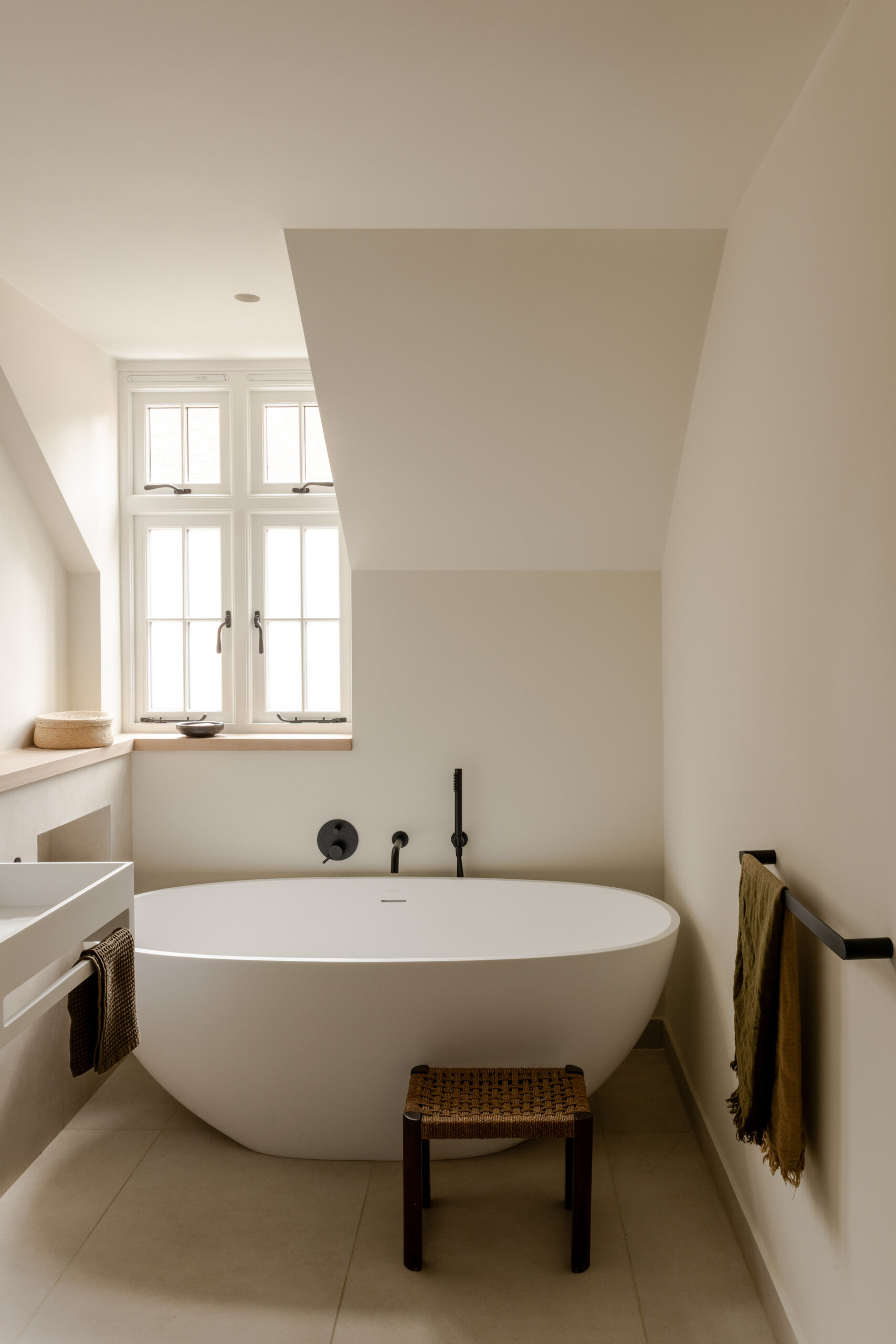 In this bathroom, a freestanding tub sits beneath a sloped ceiling, paired with warm white walls, natural wood details and slim black fixtures. A wall-mounted sink, round mirror and woven stool keep the compact room feeling open, while muted textiles add softness.