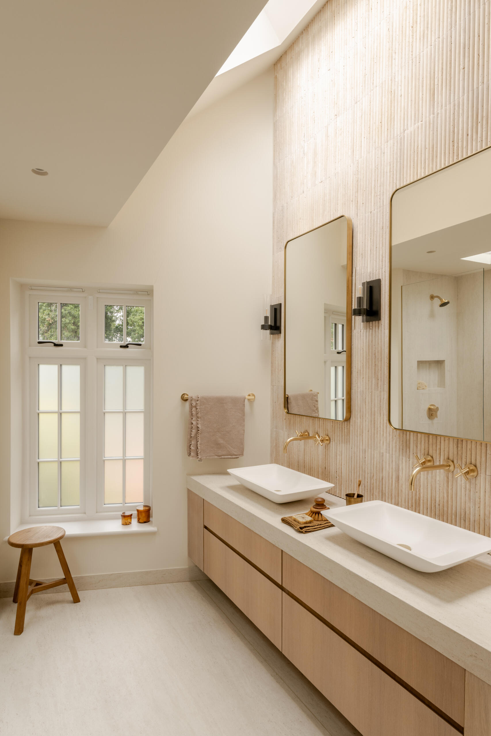 This bathroom features warm ivory travertine, pale wood cabinetry and brushed brass fixtures. A long double vanity provides generous storage, while tall mirrors and a skylight bring light deeper into the room. A frameless glass shower keeps the layout open, with a fluted wall finish, simple wood stool and muted textiles adding texture without making the space feel busy.