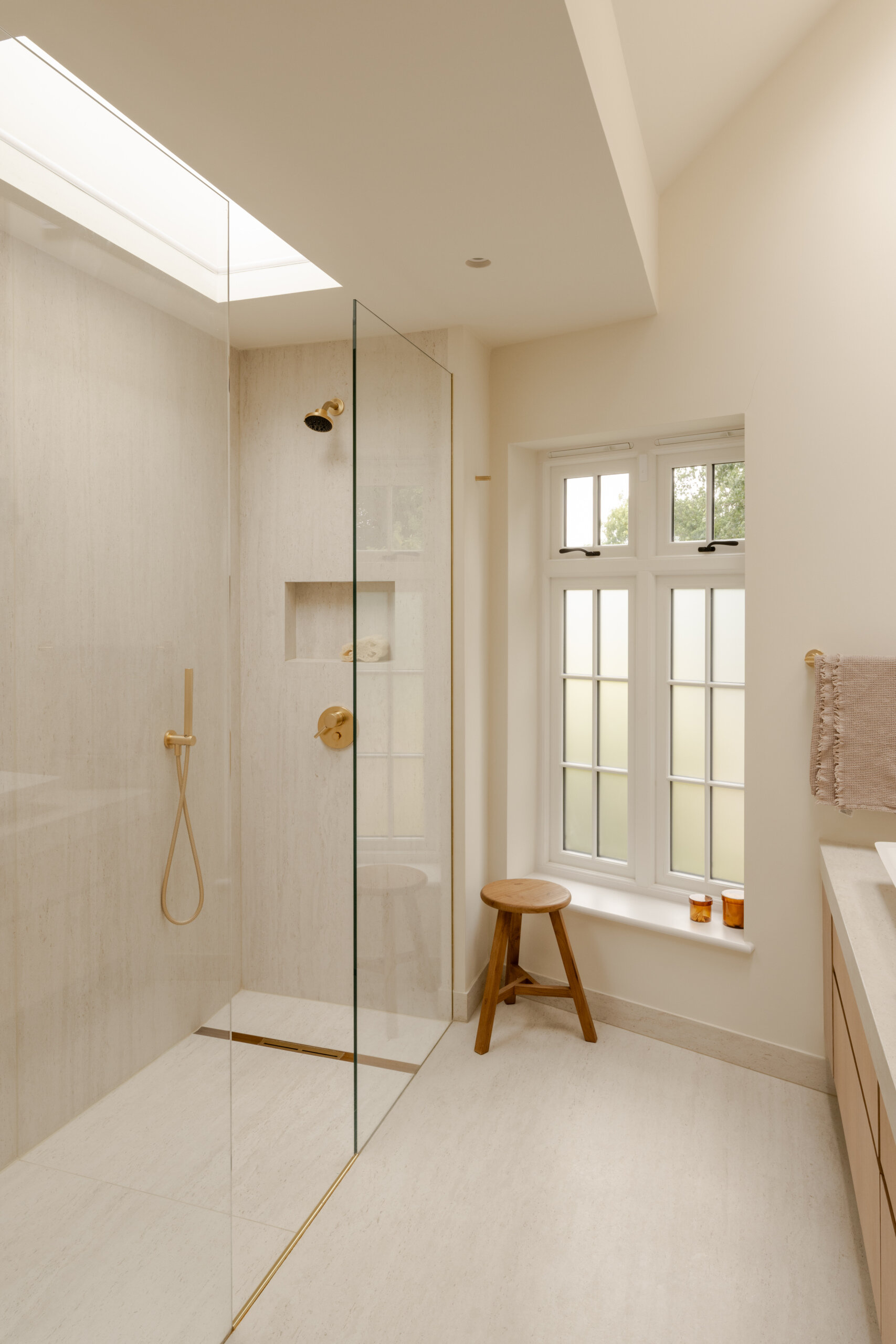 This bathroom features warm ivory travertine, pale wood cabinetry and brushed brass fixtures. A long double vanity provides generous storage, while tall mirrors and a skylight bring light deeper into the room. A frameless glass shower keeps the layout open, with a fluted wall finish, simple wood stool and muted textiles adding texture without making the space feel busy.
