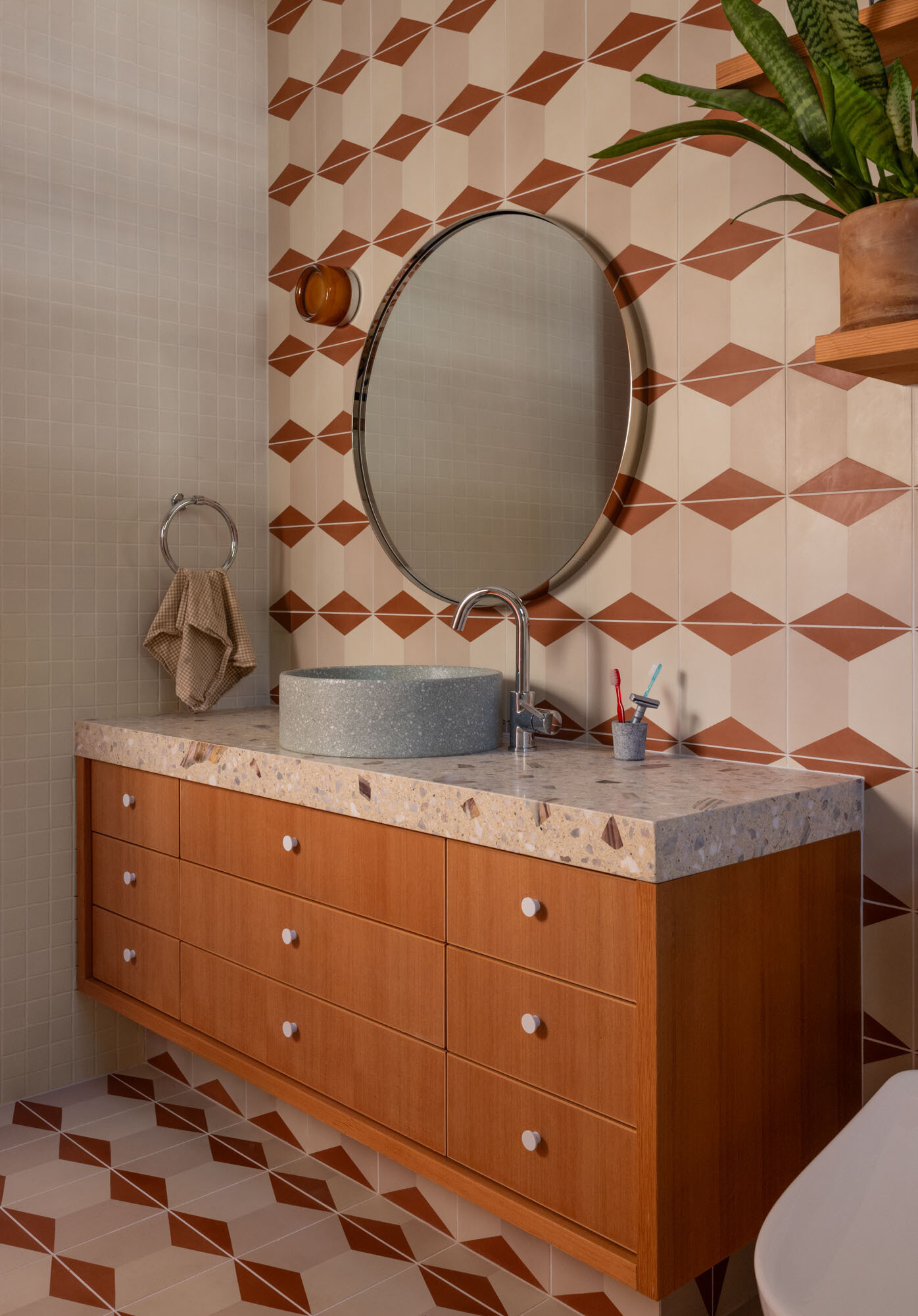 Explore this warm bathroom with cream-and-rust geometric tile, a floating timber vanity, terrazzo countertop, round vessel basin, circular mirror and simple timber shelves filled with greenery.