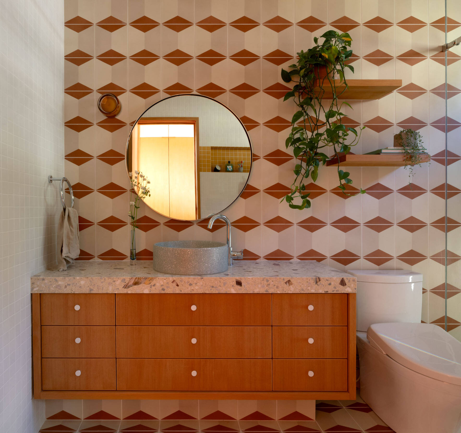 Explore this warm bathroom with cream-and-rust geometric tile, a floating timber vanity, terrazzo countertop, round vessel basin, circular mirror and simple timber shelves filled with greenery.