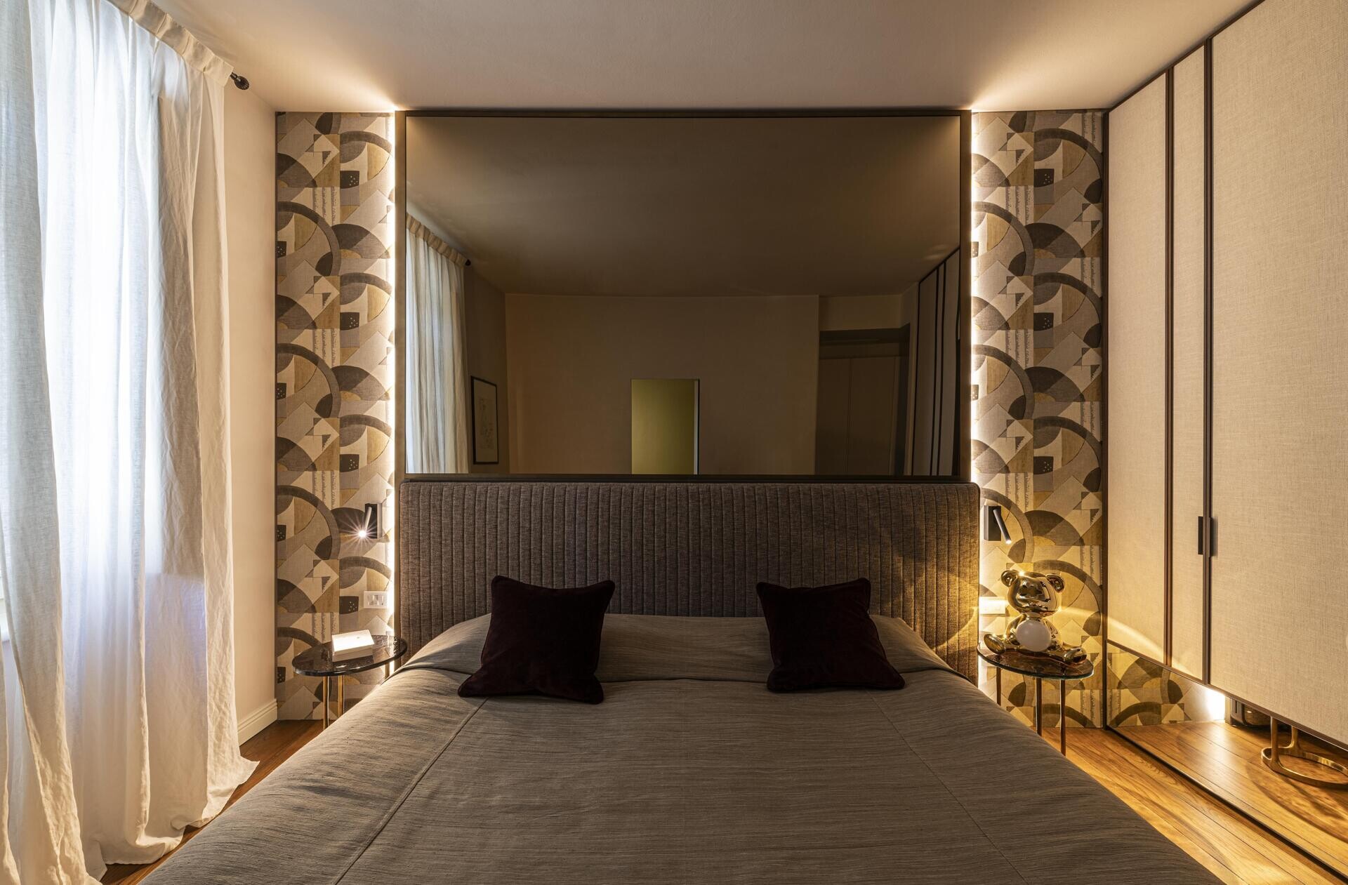 This bedroom takes a quieter, more sophisticated direction, with the bed set against a patterned wall featuring curved and geometric motifs in earthy neutrals. A mirror makes the room feel larger, while the floor-to-ceiling wardrobes in a light fabric finish keep the storage visually understated, and warm timber flooring and soft lighting add warmth.