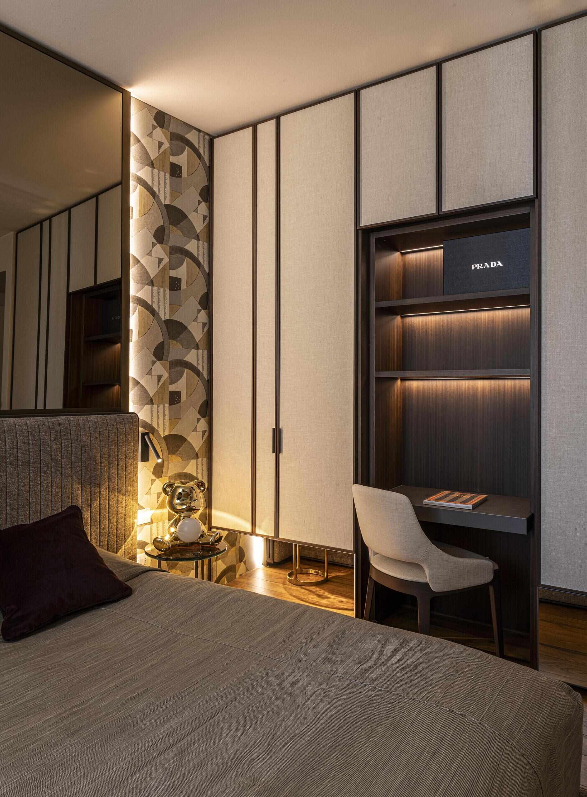 This bedroom takes a quieter, more sophisticated direction, with the bed set against a patterned wall featuring curved and geometric motifs in earthy neutrals. A mirror makes the room feel larger, while the floor-to-ceiling wardrobes in a light fabric finish keep the storage visually understated, and warm timber flooring and soft lighting add warmth.