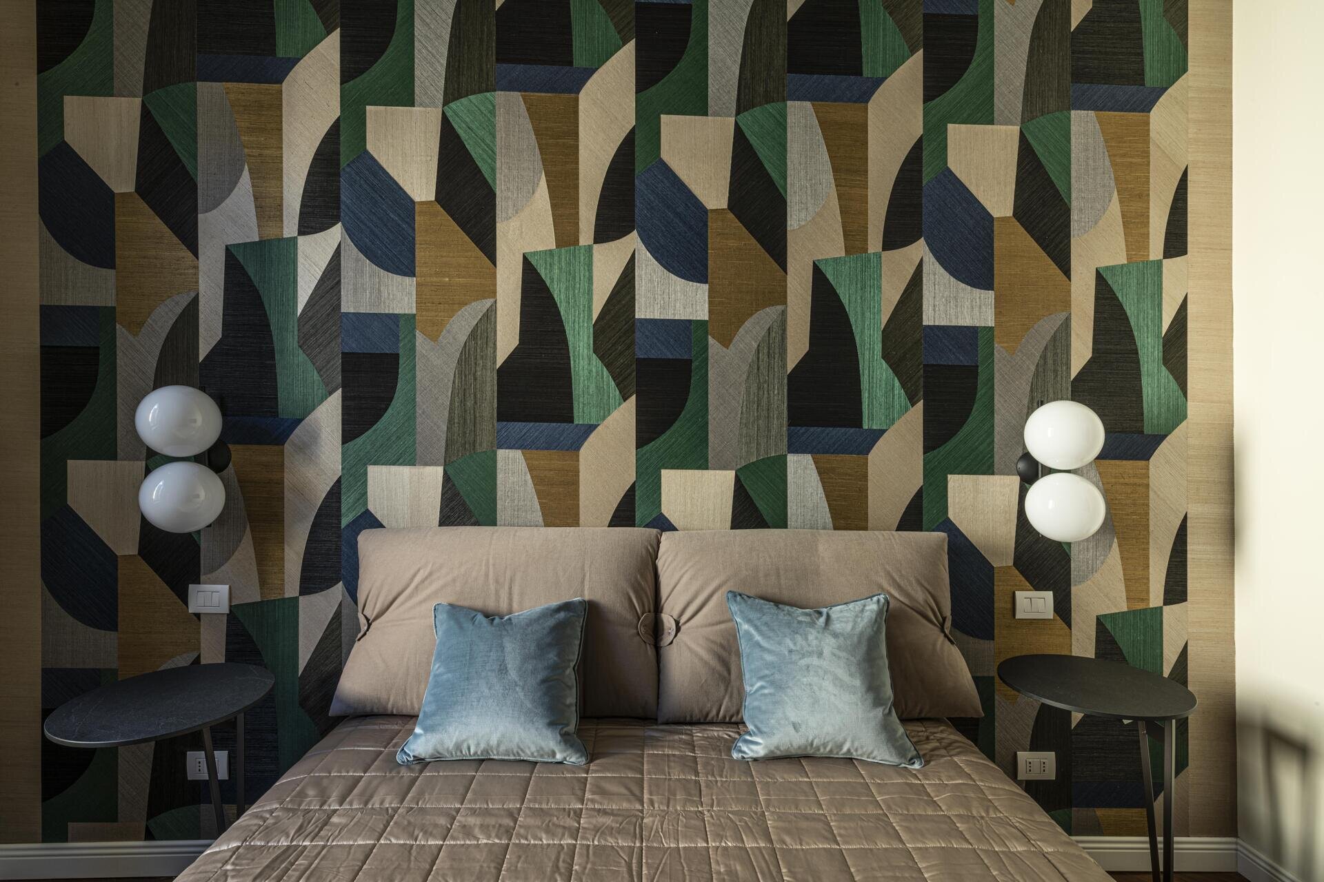 This bedroom pairs an upholstered headboard with a dramatic geometric wall covering in deep green, navy, taupe and warm brown. The pattern stretches across the wall behind the bed, framed by simple black side tables and sculptural globe lamps, while soft blue cushions add another layer of colour.