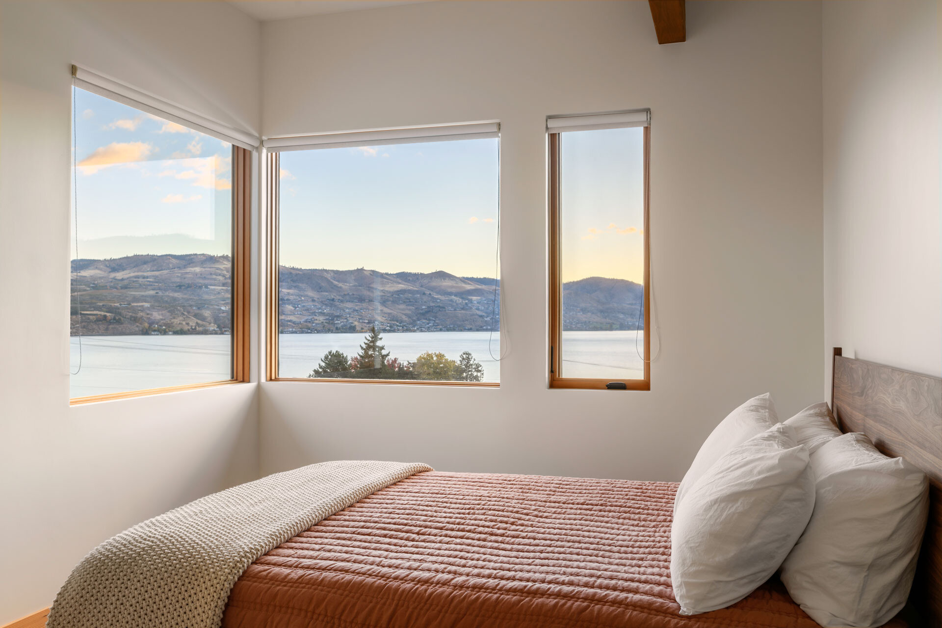 A serene guest bedroom features soft white walls, wood-framed windows, a rust-coloured quilt, and sweeping lake and mountain views.