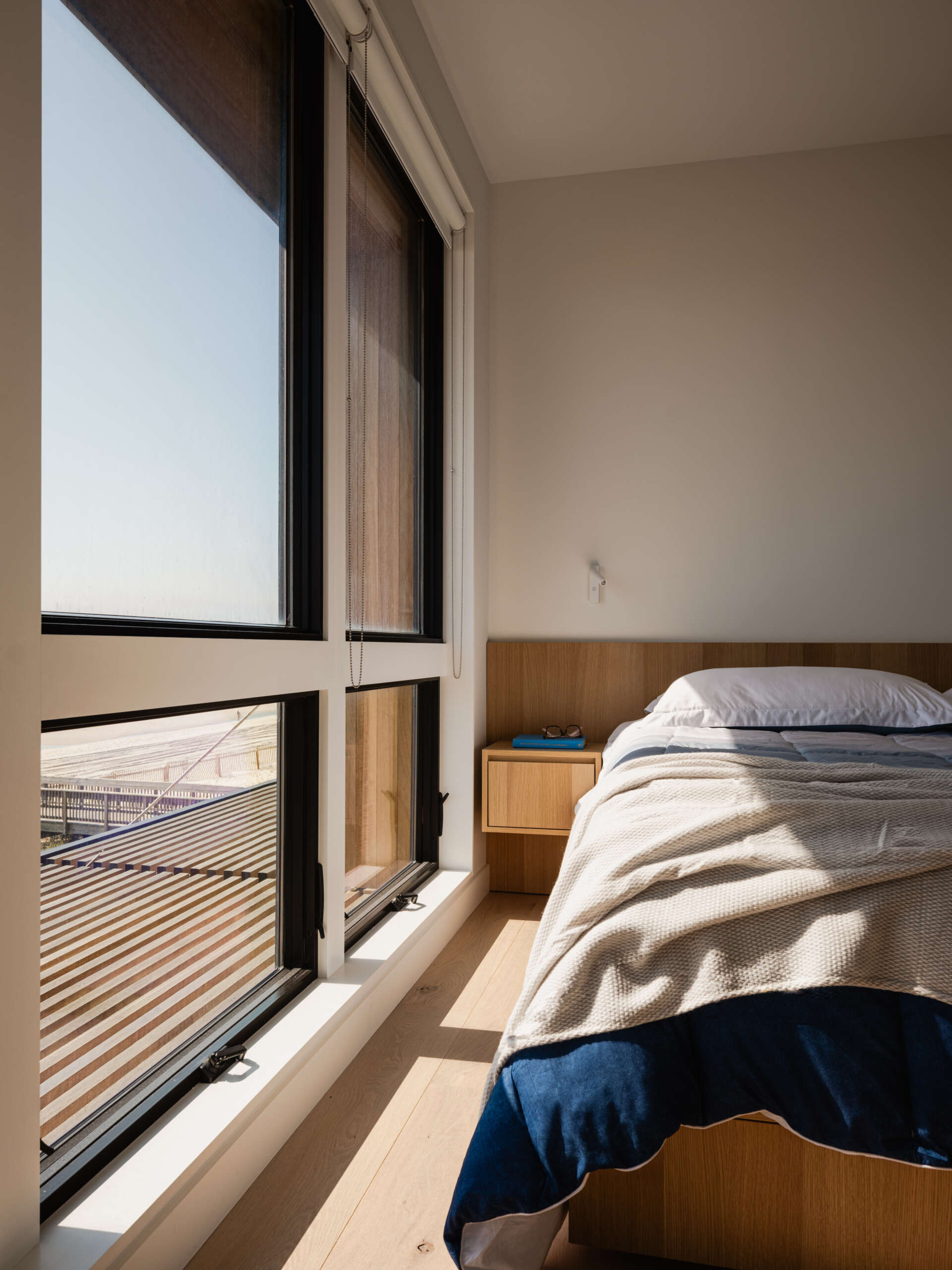 Tall windows, a wood headboard, and a floating bedside drawer give this Fire Island bedroom a simple coastal style. Blue and cream bedding complements the pale wood floor, with beach glimpses just outside.