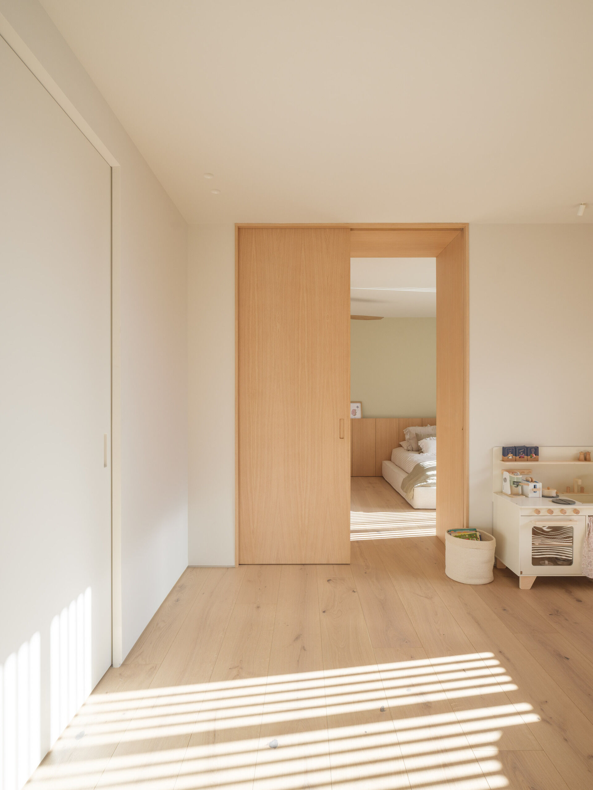 In the upstairs bedroom, a large dark-framed window looks across the planted roof, bringing grasses and trees into view from the bed. White bedding and an upholstered headboard are layered with brown and cream cushions, alongside simple wooden bedside drawers. Small black pendant lights hang on either side, leaving the bedside surfaces free, while a ceiling fan with curved wooden blades echoes the timber floor below.