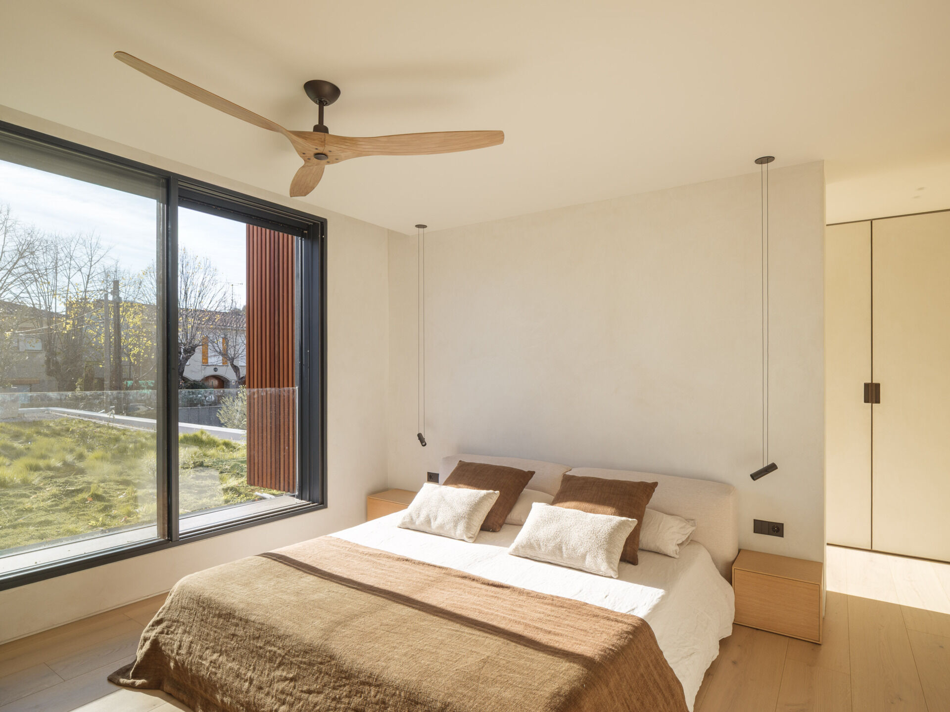 In the upstairs bedroom, a large dark-framed window looks across the planted roof, bringing grasses and trees into view from the bed. White bedding and an upholstered headboard are layered with brown and cream cushions, alongside simple wooden bedside drawers. Small black pendant lights hang on either side, leaving the bedside surfaces free, while a ceiling fan with curved wooden blades echoes the timber floor below.