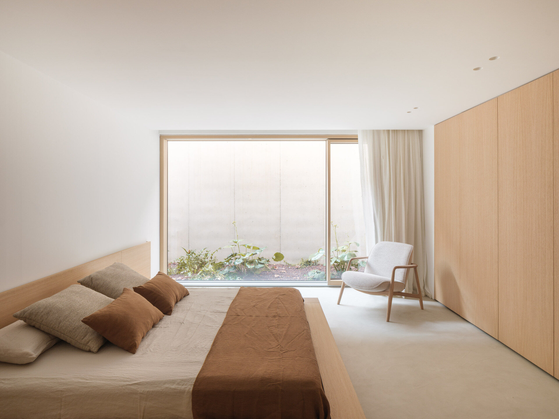 This bedroom has an enclosed outlook, with wide glazing facing a planted courtyard and a tall pale wall. A low timber bed sits opposite full-height wooden wardrobes, paired with neutral bedding and a cinnamon-coloured throw. An upholstered armchair beside the window offers a spot to read, and a sheer curtain softens the edge of the glass. Here, the courtyard supplies daylight and a close view of greenery while keeping the room sheltered from neighbouring views.