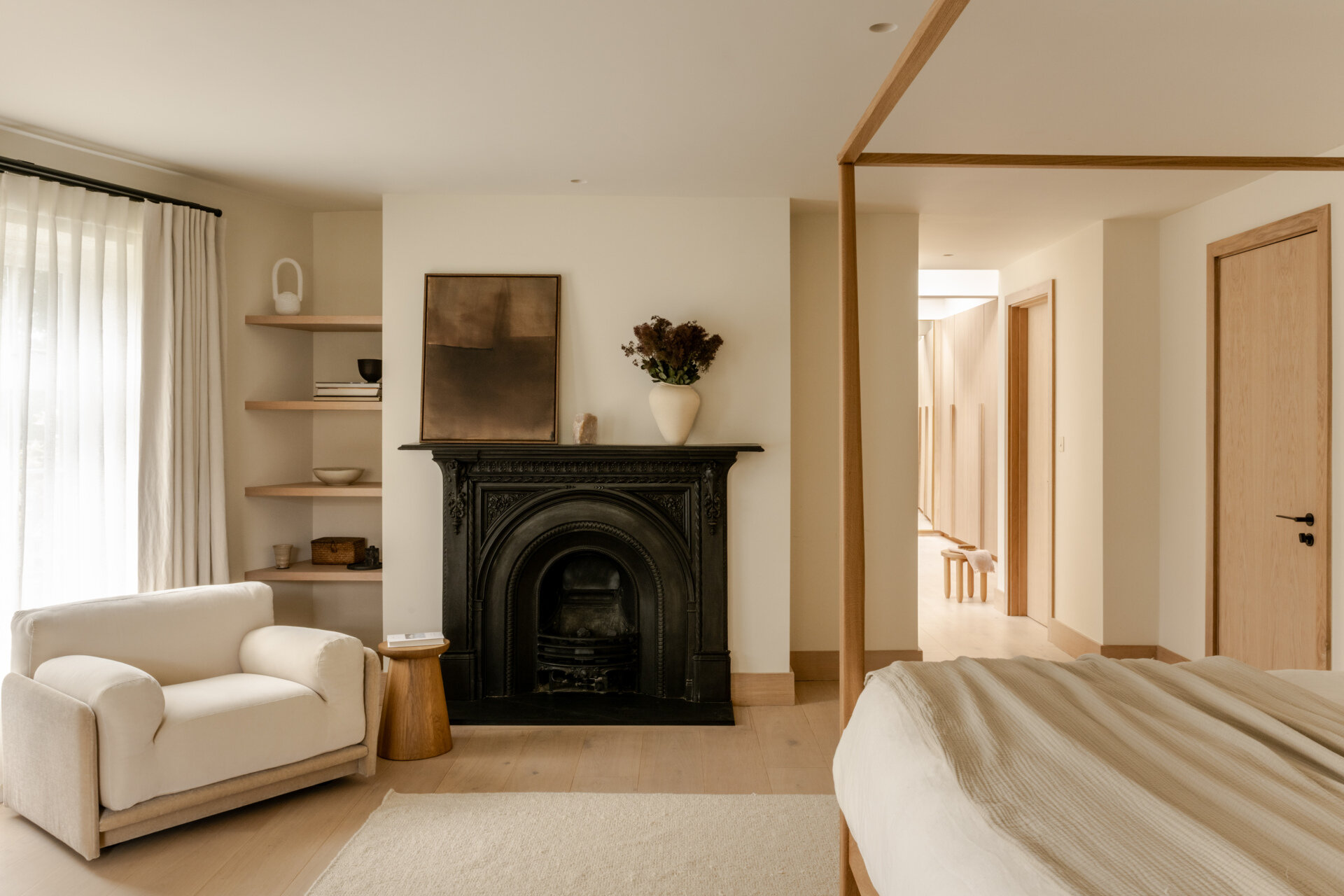Step inside the primary bedroom at Thirsk Cottage, featuring soft neutral tones, pale oak flooring, warm wood, an ornate black fireplace and a slim four-poster bed.
