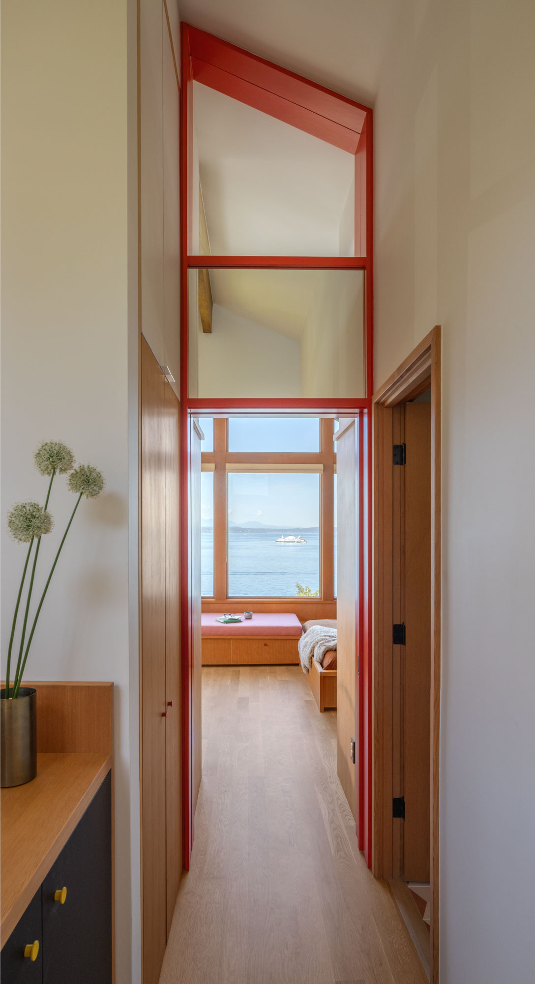 Explore the updated upper hallway, where pale timber flooring and a tall red-framed glazed transom pull light deeper into the house and create a clear view toward the bedroom and Puget Sound beyond.