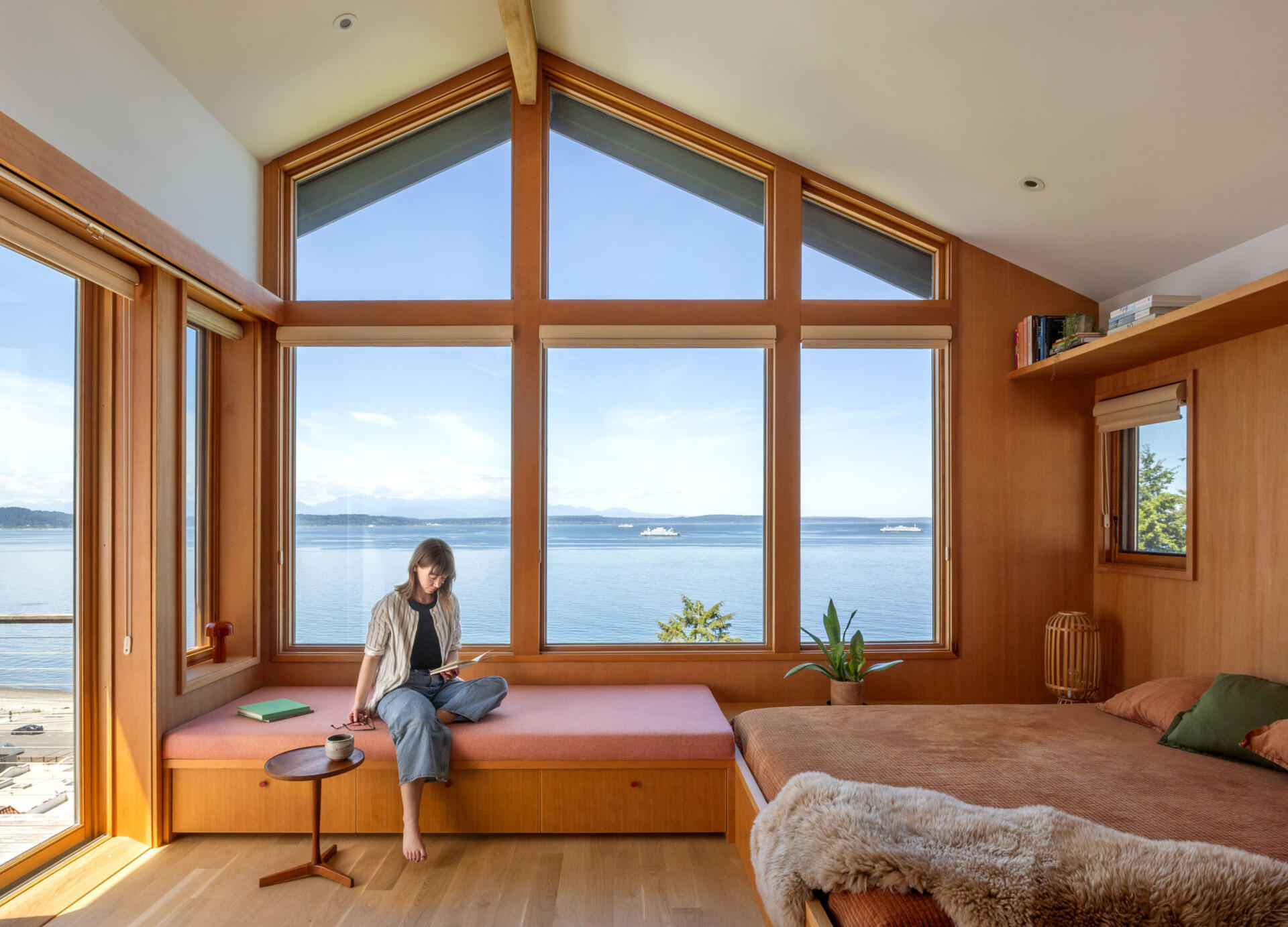 Tour the primary bedroom at Alki Lookout, where SHED paired existing panoramic Puget Sound windows with pale timber built-ins, a custom bed platform, concealed drawers and a pink-cushioned window bench.