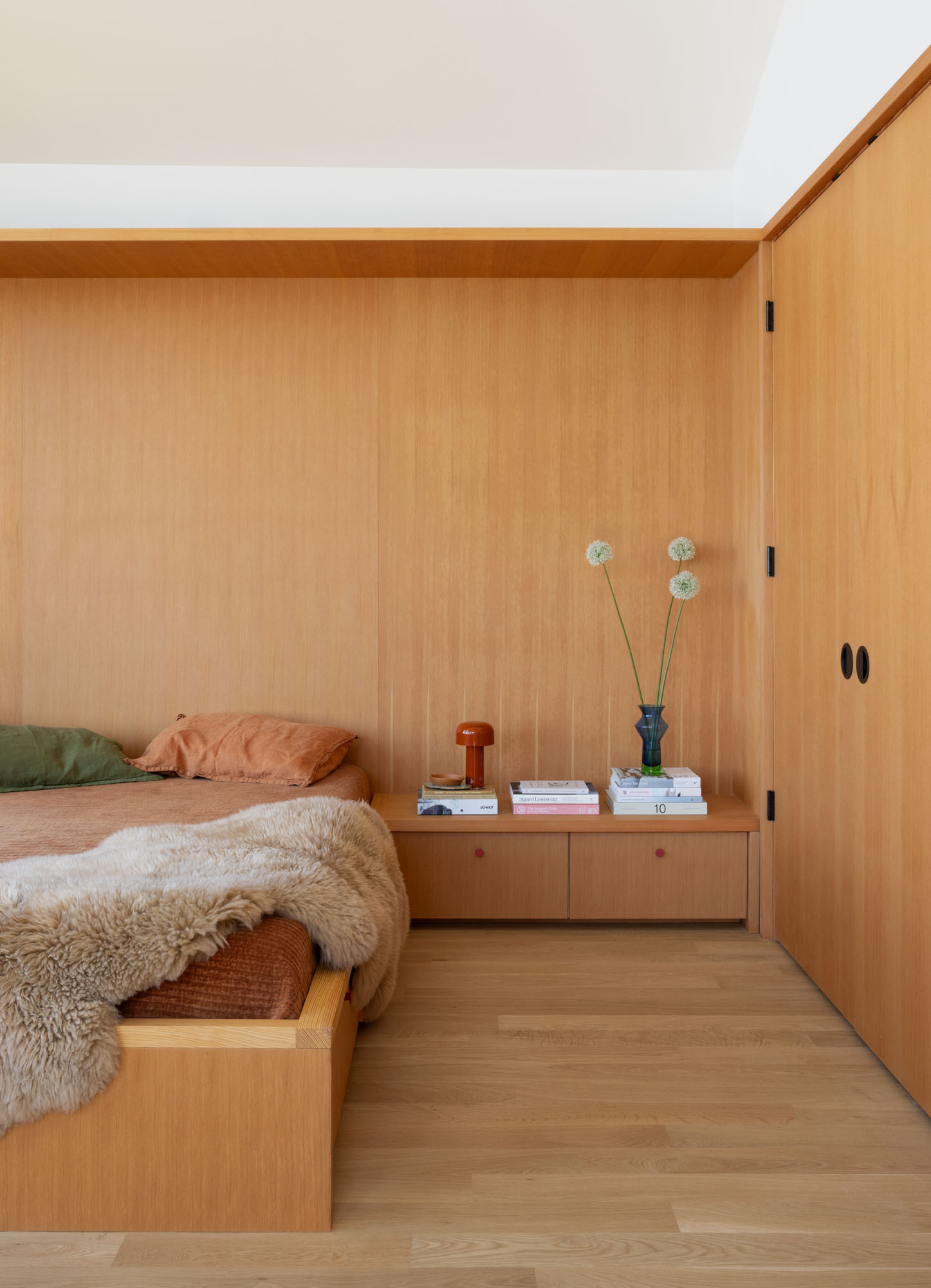 Tour the primary bedroom at Alki Lookout, where SHED paired existing panoramic Puget Sound windows with pale timber built-ins, a custom bed platform, concealed drawers and a pink-cushioned window bench.