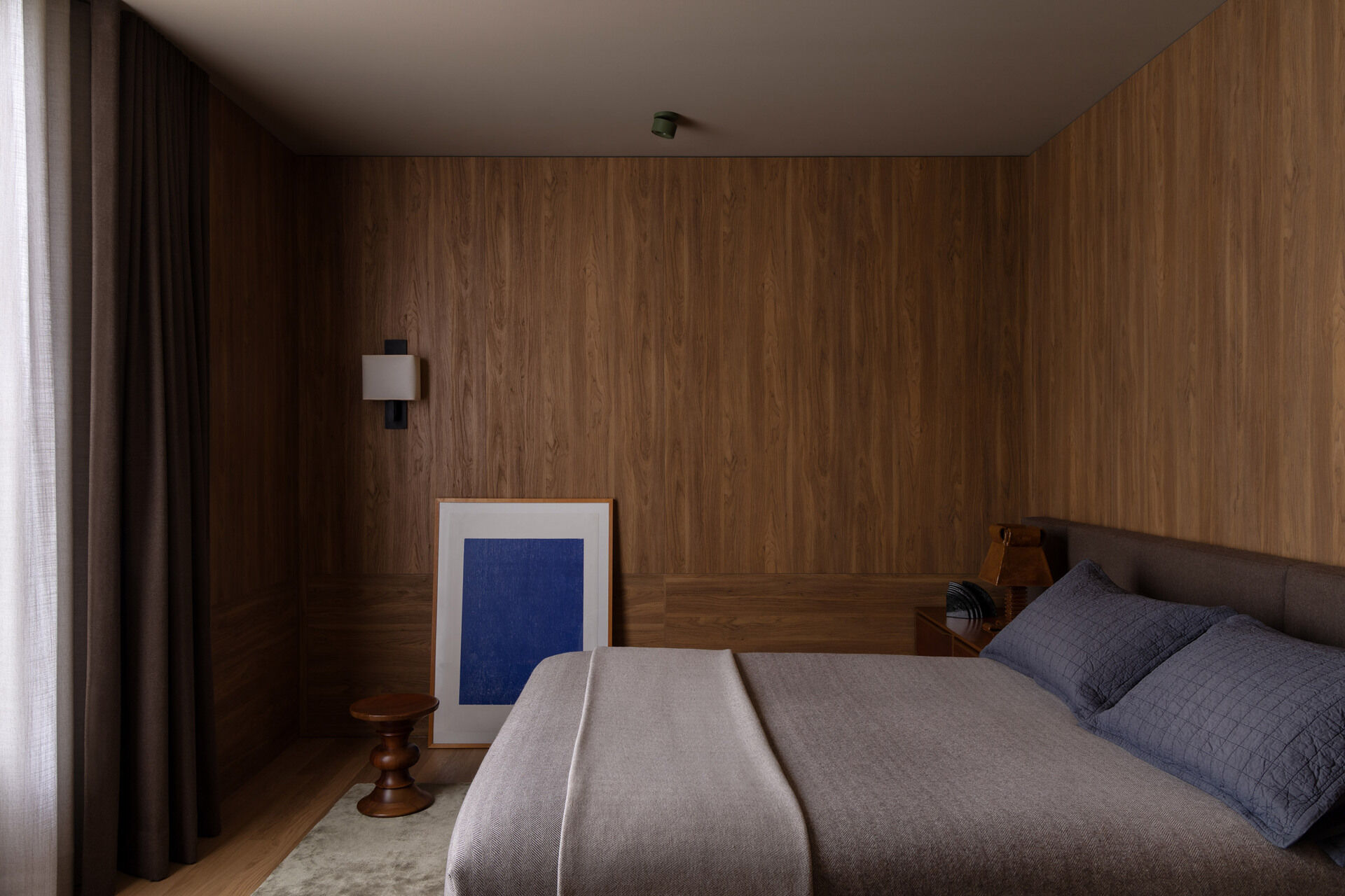 This bedroom continues the house’s use of timber, with warm oak lining the walls and built-in cabinetry extending into the adjoining dressing area. The bed sits against a dark upholstered headboard, while soft grey bedding, sheer curtains and a small framed artwork keep the room subdued.