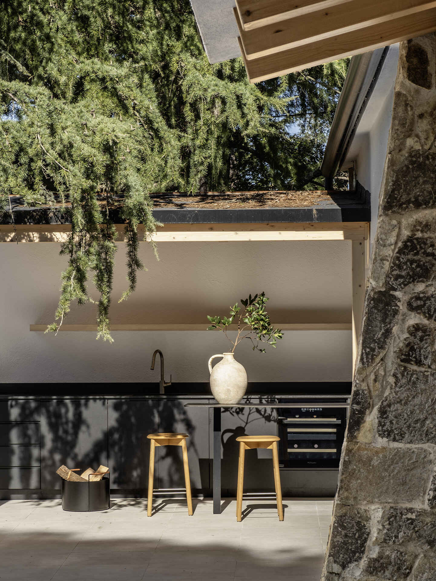 The outdoor spaces include a dark contemporary kitchen, a broad pool terrace and a barrel-shaped timber sauna tucked between mature trees. Together, they create several places for cooking, swimming and relaxing throughout the garden.