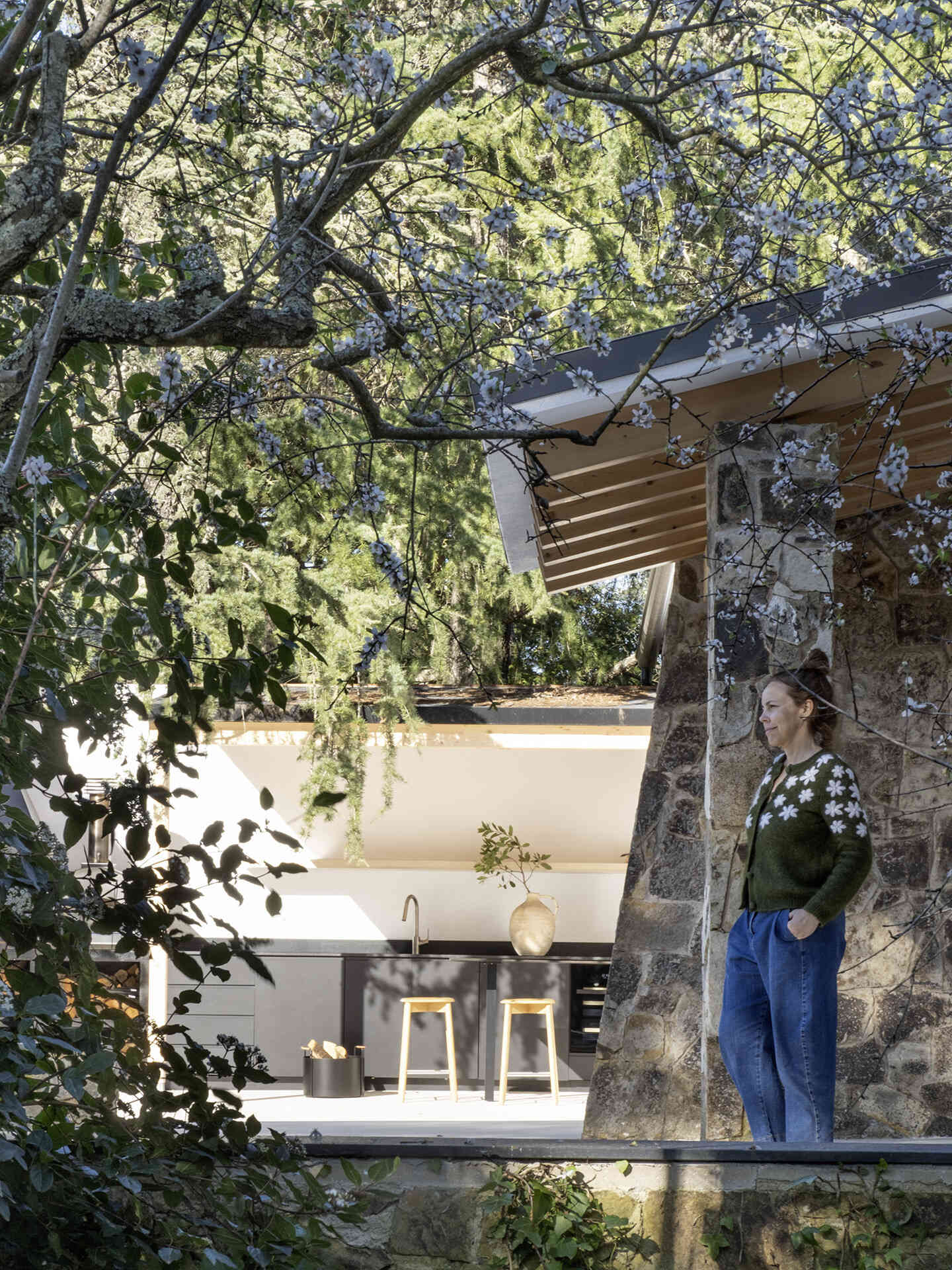 The outdoor spaces include a dark contemporary kitchen, a broad pool terrace and a barrel-shaped timber sauna tucked between mature trees. Together, they create several places for cooking, swimming and relaxing throughout the garden.