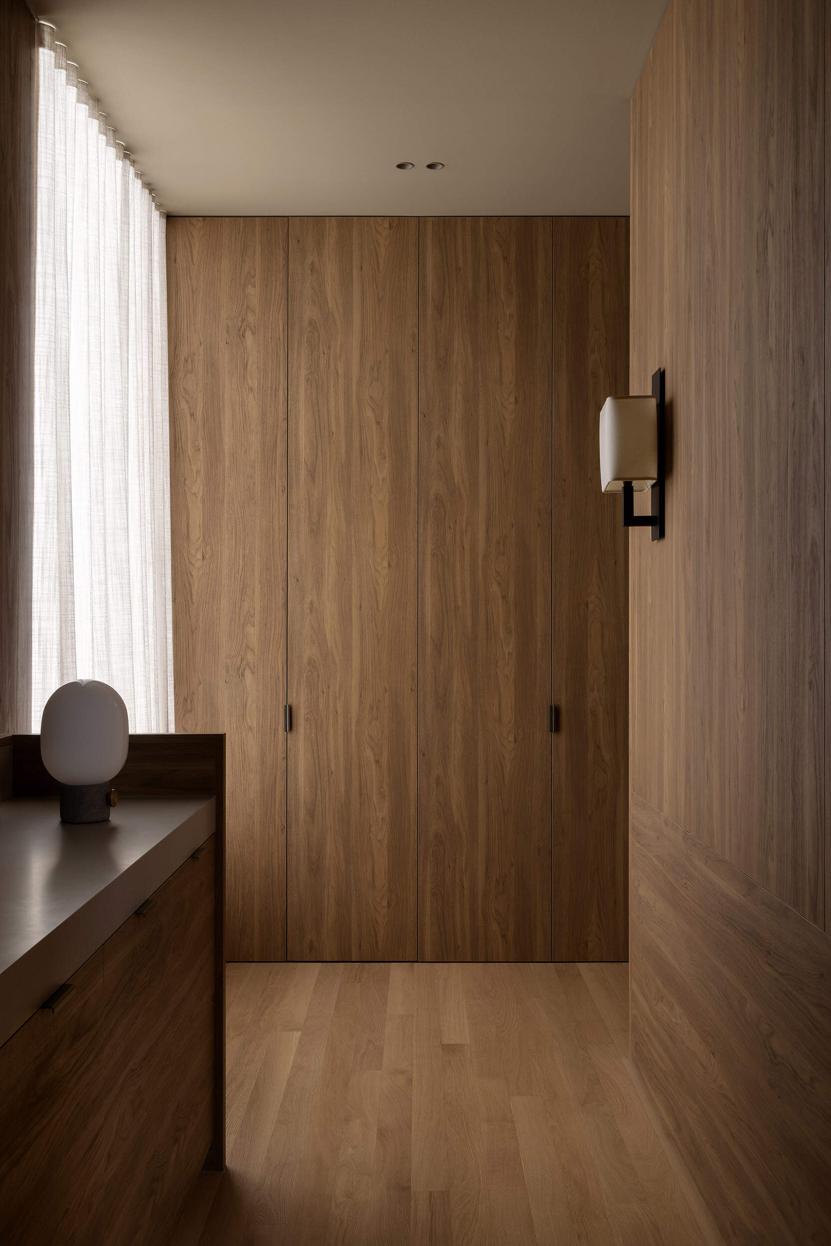 In this closet, full-height doors conceal storage, with a timber vanity and simple wall light continuing the same furniture-like approach to joinery seen downstairs.
