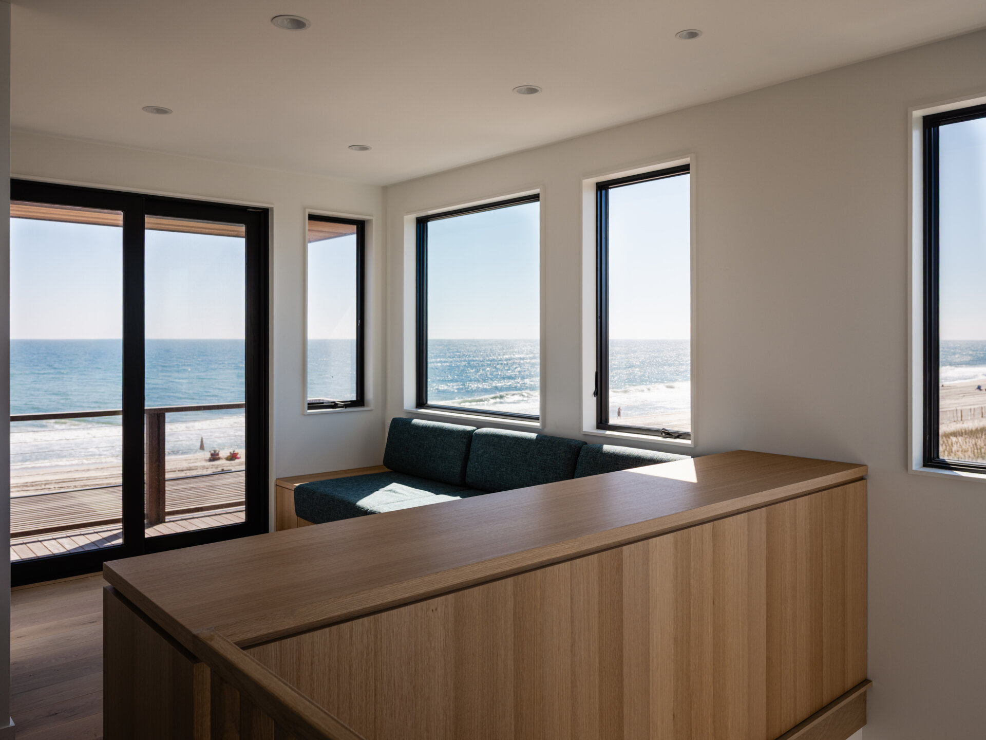 A built-in cushioned seat transforms this upstairs landing into a compact sitting area overlooking the ocean. Corner windows and balcony access make the most of the beach views at Surf Road House on Fire Island.