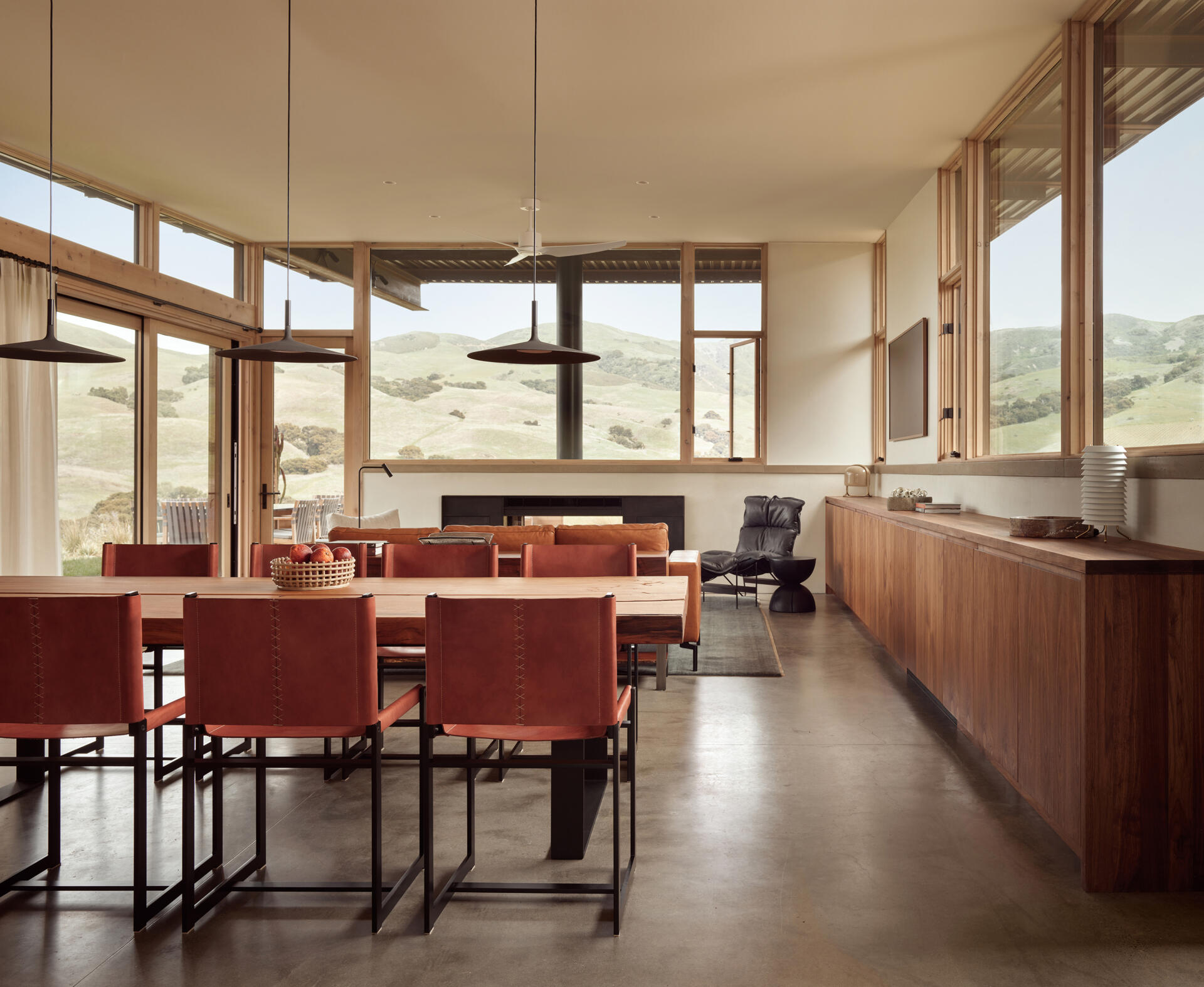 The dining area sits within the open-plan great room, positioned between the living spaces and the kitchen. A long wooden dining table takes center stage, surrounded by leather-upholstered chairs with slim black frames. Above it, three simple pendant lights hang in a row, adding a clean architectural detail over the table.