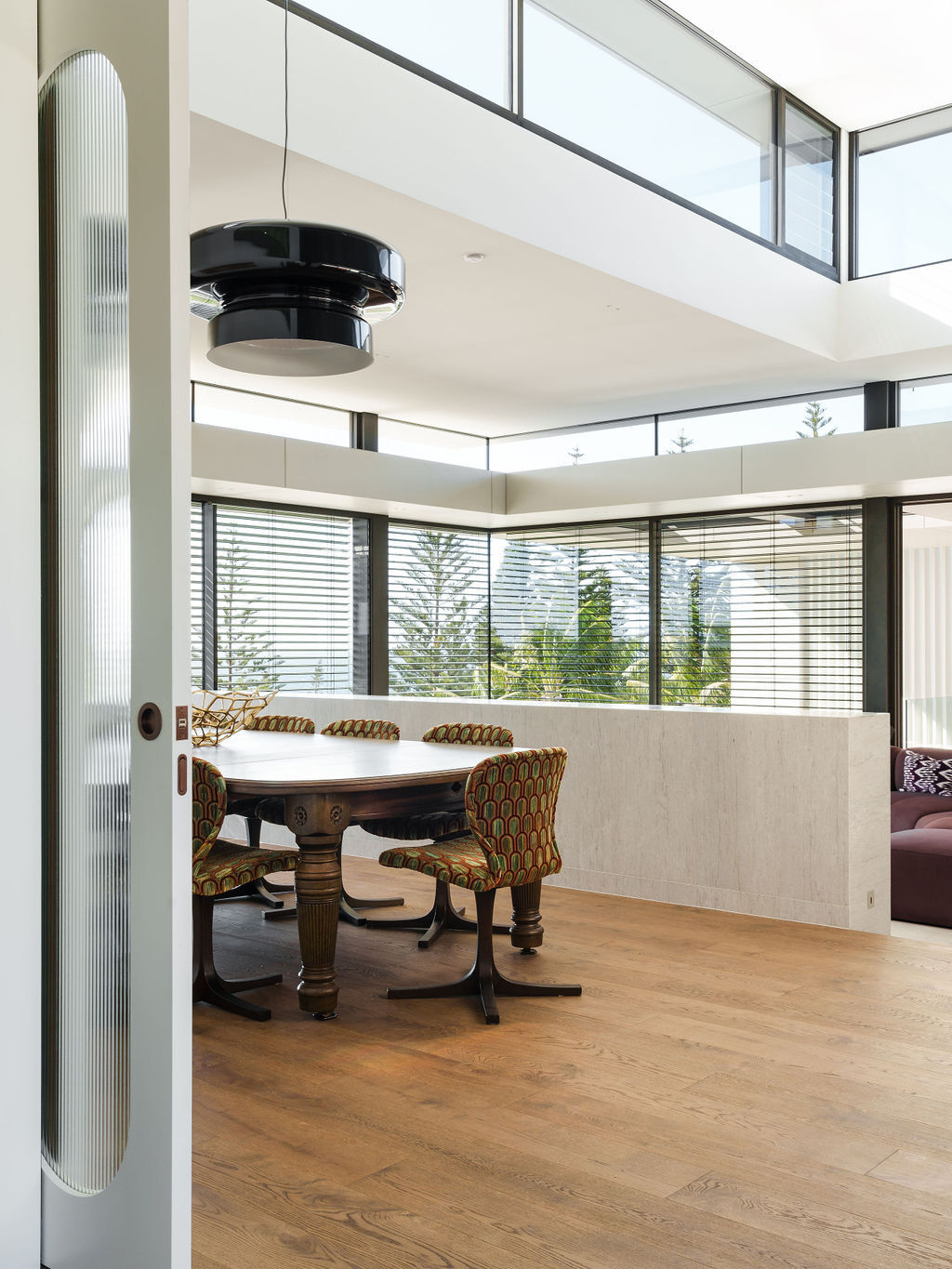 A double-height dining room at Mezzanine House pairs a round timber table, patterned chairs and a sculptural black pendant with expansive glazing and treetop views.