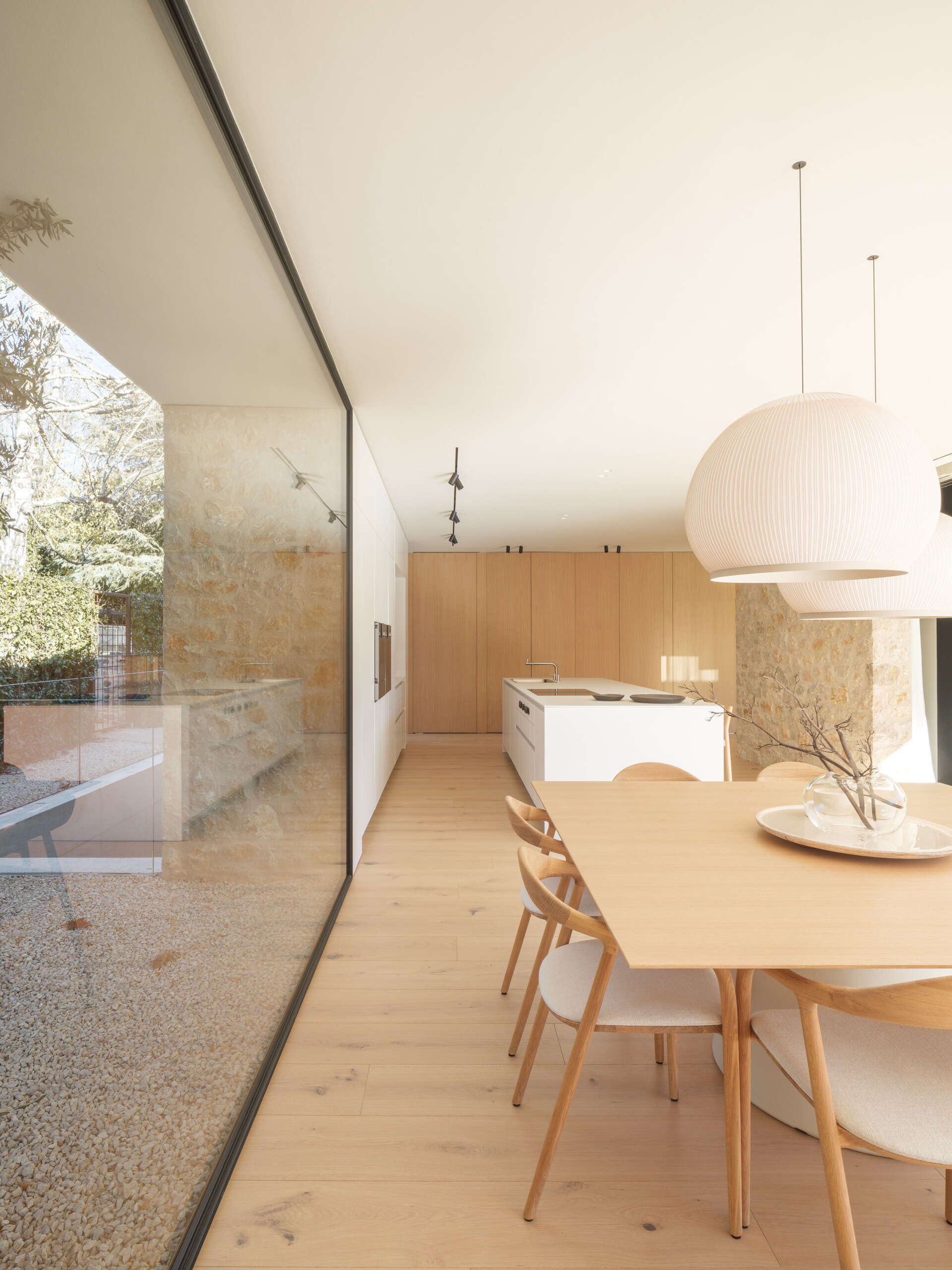 A timber dining table and rounded white pendants sit between garden views and a tree-filled courtyard. Explore the adjoining kitchen, with a white island, full-height wooden cabinetry, and exposed stone at House 2194 in Olot, Spain.