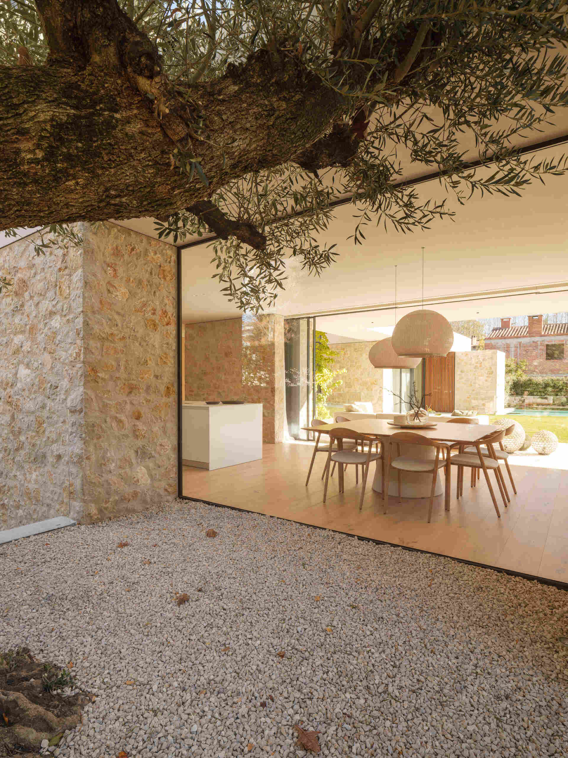 A timber dining table and rounded white pendants sit between garden views and a tree-filled courtyard. Explore the adjoining kitchen, with a white island, full-height wooden cabinetry, and exposed stone at House 2194 in Olot, Spain.