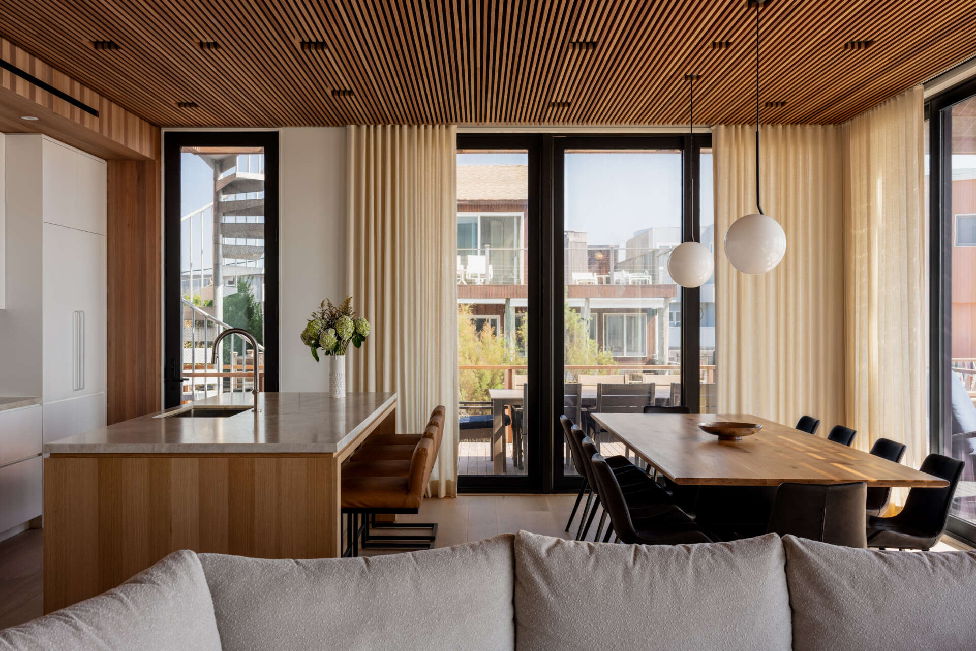 White cabinetry, a wood kitchen island, and tan counter stools meet a long dining table in this open-plan Fire Island home. Globe pendants and trellis-filtered sunlight add character, with sliding doors leading to outdoor dining.