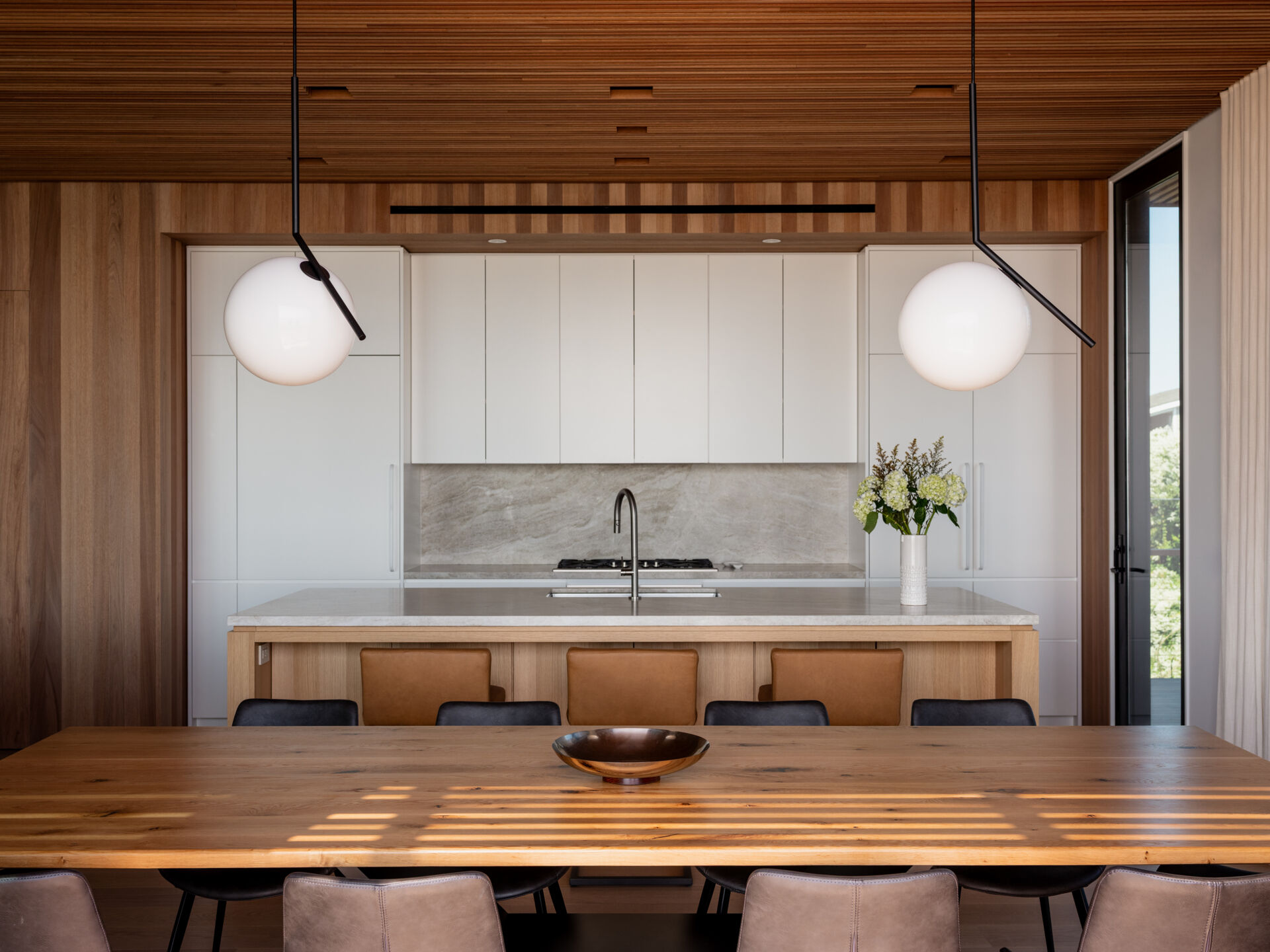 White cabinetry, a wood kitchen island, and tan counter stools meet a long dining table in this open-plan Fire Island home. Globe pendants and trellis-filtered sunlight add character, with sliding doors leading to outdoor dining.