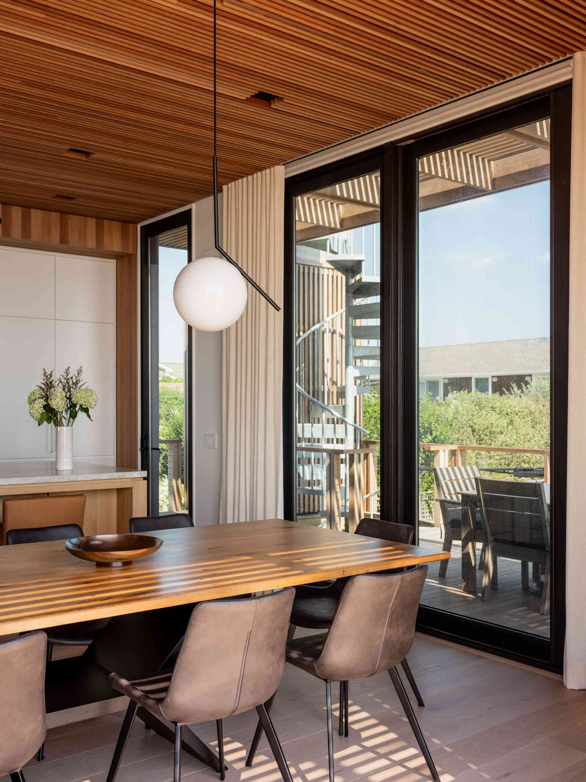 White cabinetry, a wood kitchen island, and tan counter stools meet a long dining table in this open-plan Fire Island home. Globe pendants and trellis-filtered sunlight add character, with sliding doors leading to outdoor dining.